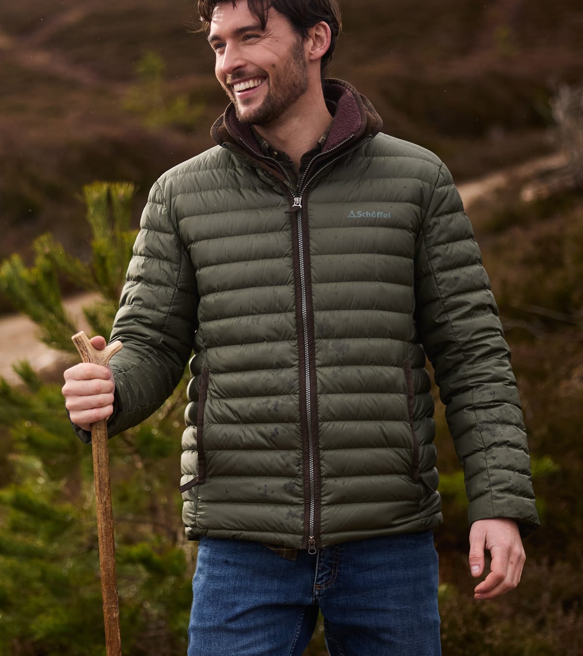 A man wearing an green puffer jacket with a dark brown zipper standing outdoors holding a wooden stick.