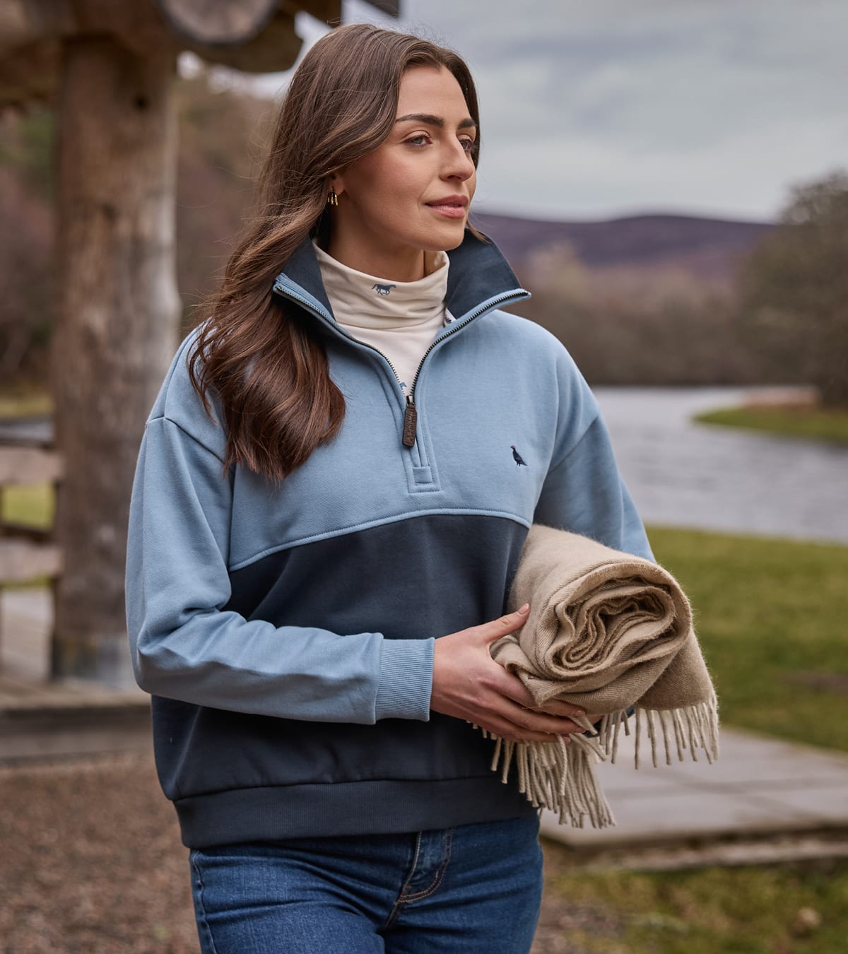 A woman outdoors near a river, wearing a colour-blocked quarter-zip sweatshirt in light blue and navy.