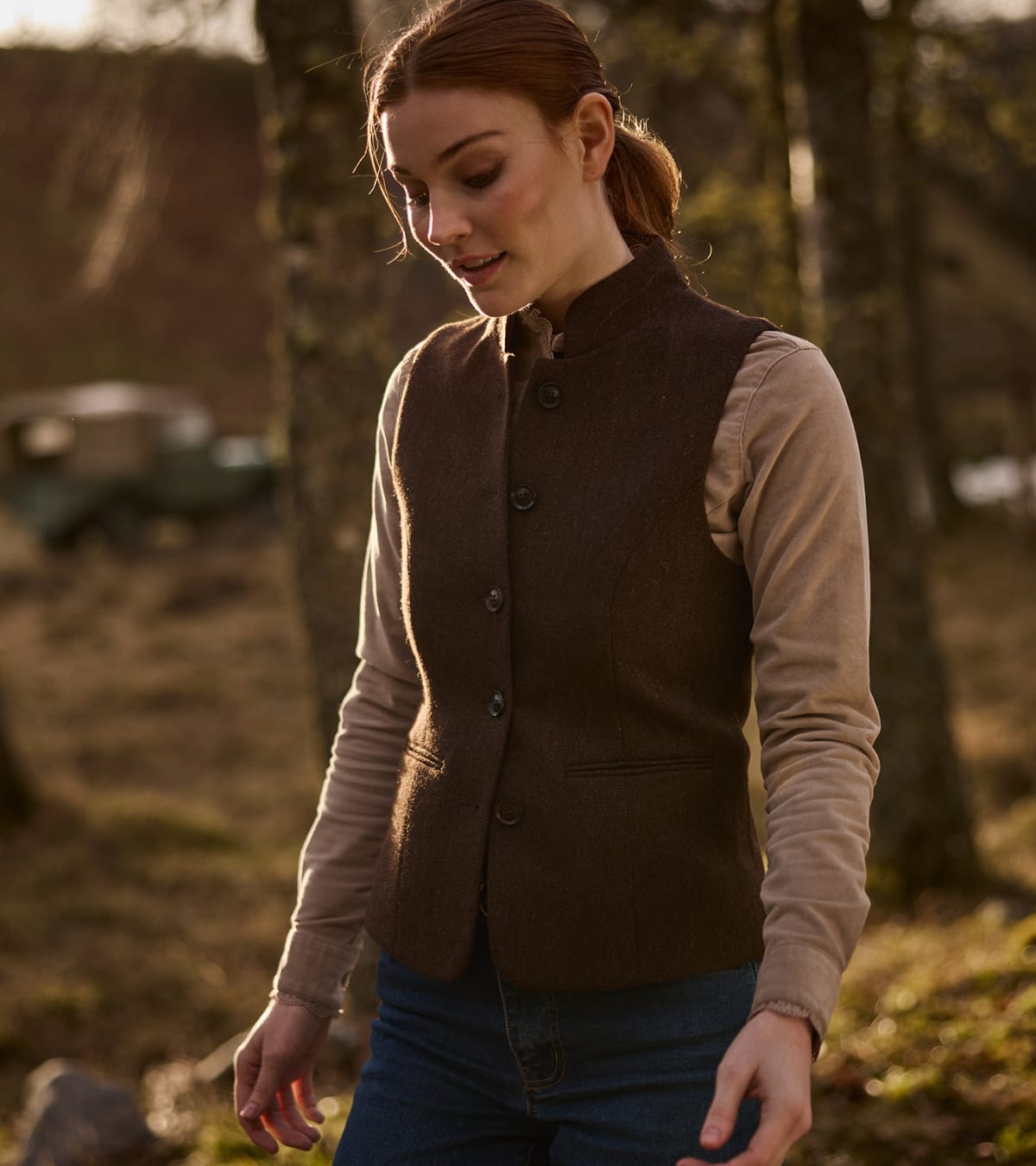 A woman wearing a dark brown tweed waistcoat standing outdoors in a wooded setting.