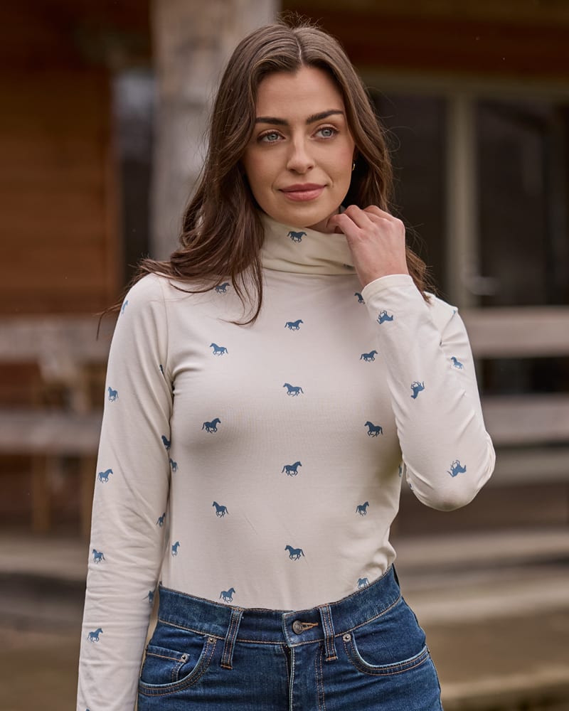 A woman wearing a cream-colored long-sleeve mock neck top with a small blue horse pattern.