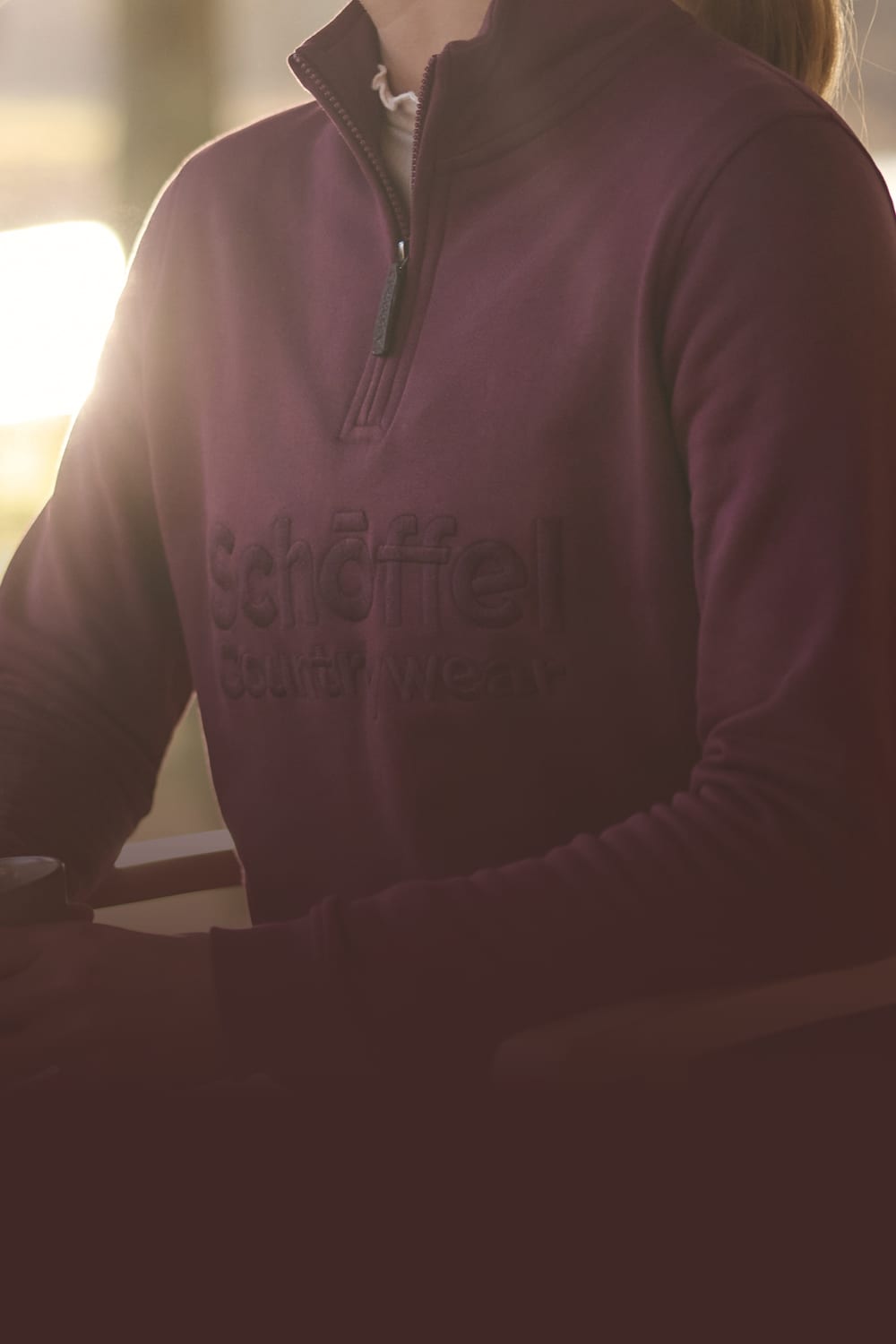 A person wearing a dark plum quarter-zip sweatshirt with embroidered 'Schöffel Countrywear' logo