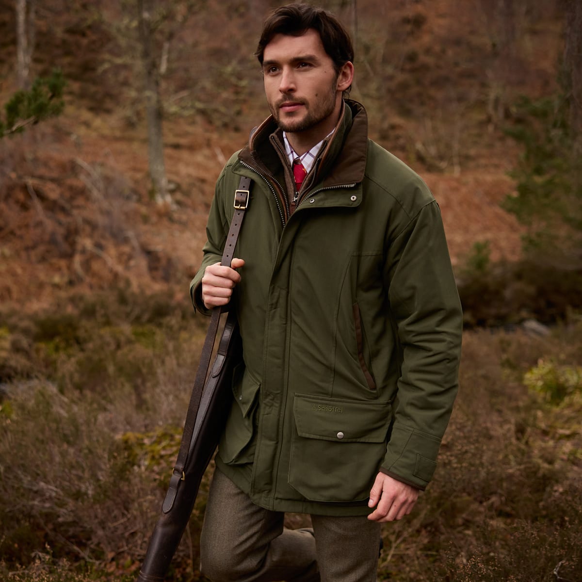 A man wearing an olive green shooting jacket with brown lining, standing in a wooded outdoor setting.