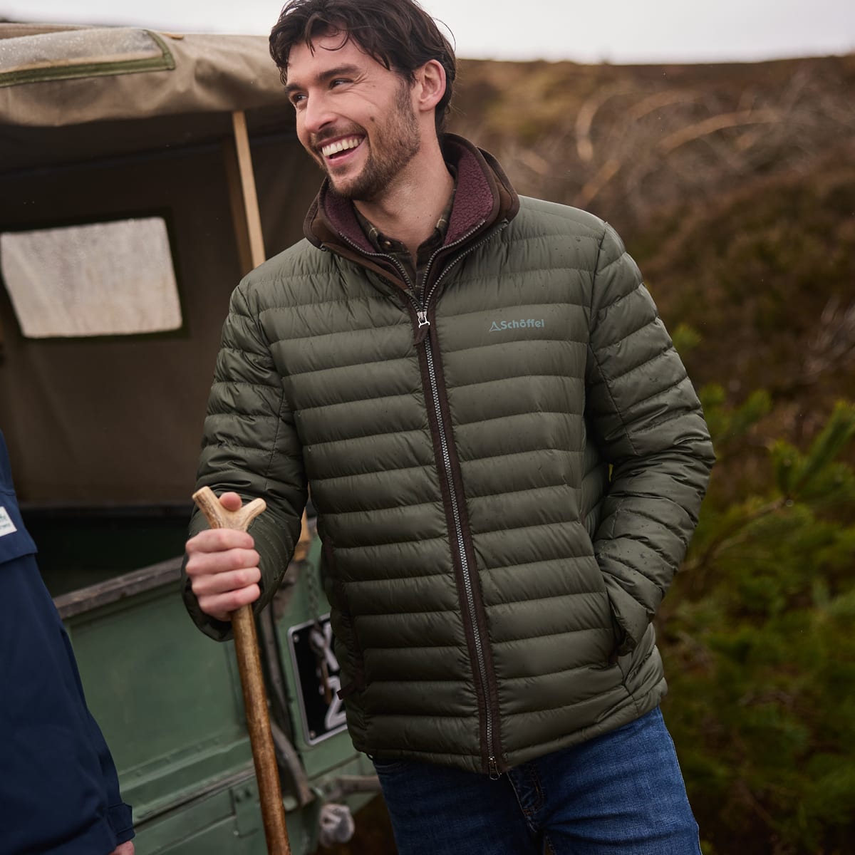 A man outdoors wearing a green puffer jacket, holding a wooden walking stick next to a vintage green vehicle.