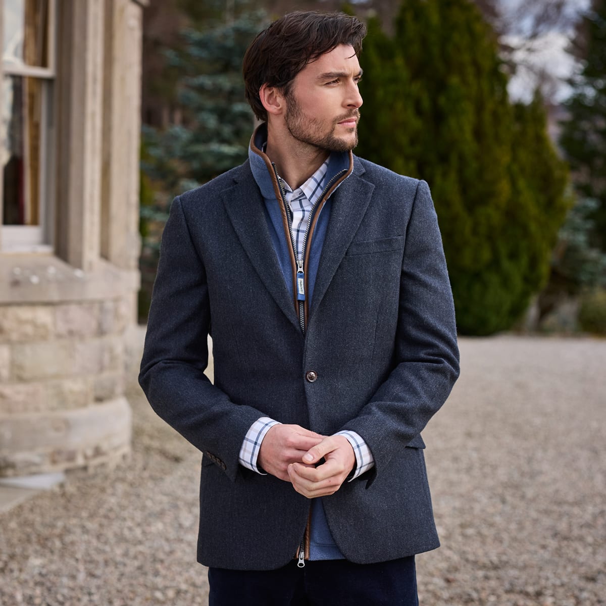 A man wearing a navy tweed blazer standing outdoors in front of a stone building and green trees.
