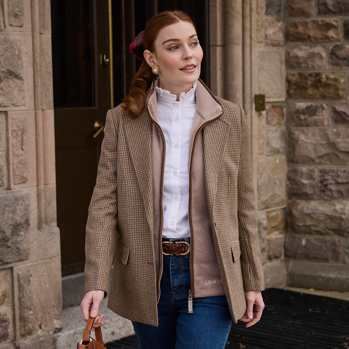 A woman wearing a tweed blazer over a white ruffled blouse, and blue jeans, standing in front of a stone building entrance.