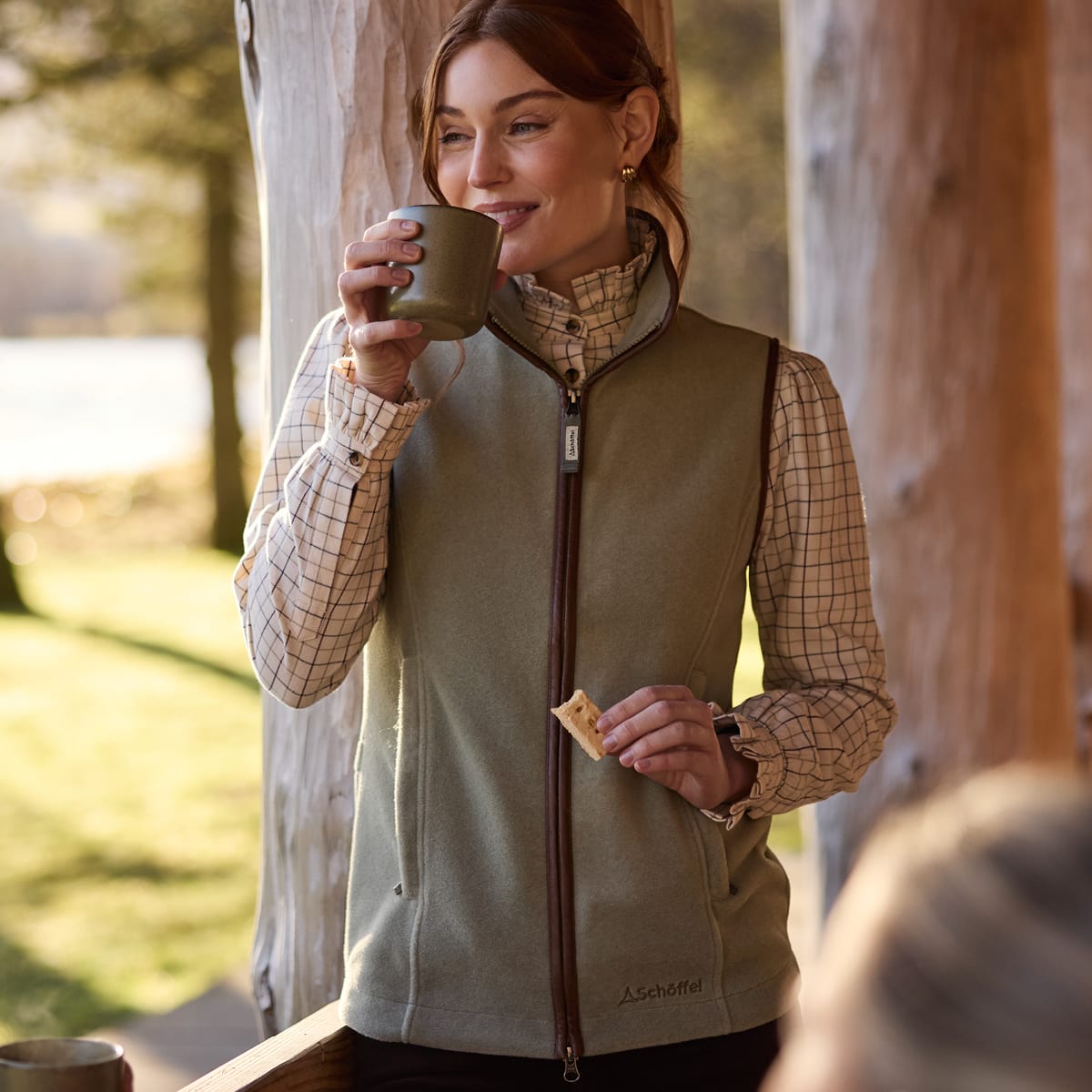 Woman drinking from a mug wearing a sage green fleece gilet over a checked shirt