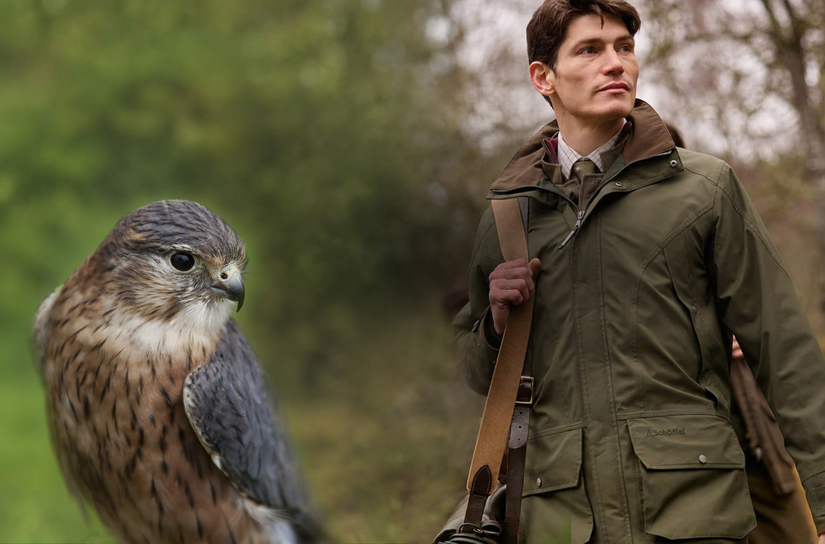 A man in an olive green hunting jacket stands outdoors with a large falcon next to him