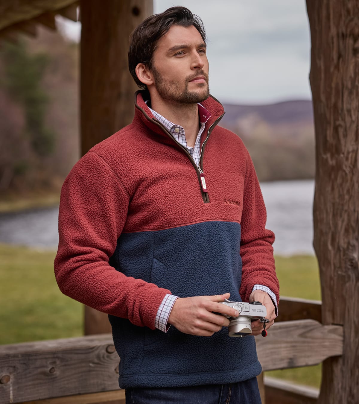 Man wearing a red and navy two-tone fleece pullover outdoors by a lake with hills in the background.