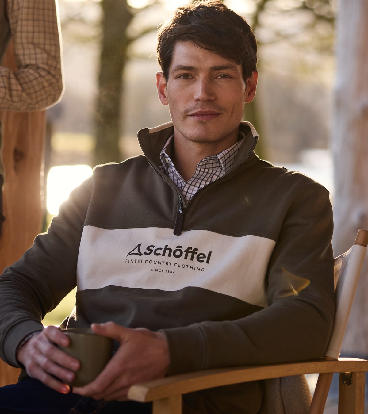 Man wearing a brown and cream Schöffel quarter-zip pullover over a checked shirt, holding a mug outdoors.