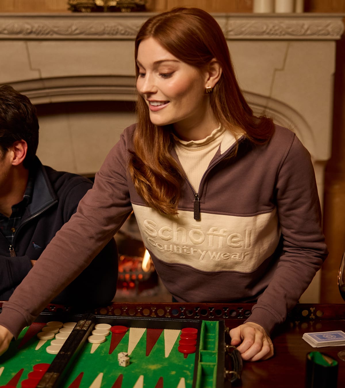 Woman wearing a brown and beige colour-block Schöffel quarter-zip pullover while playing backgammon indoors.