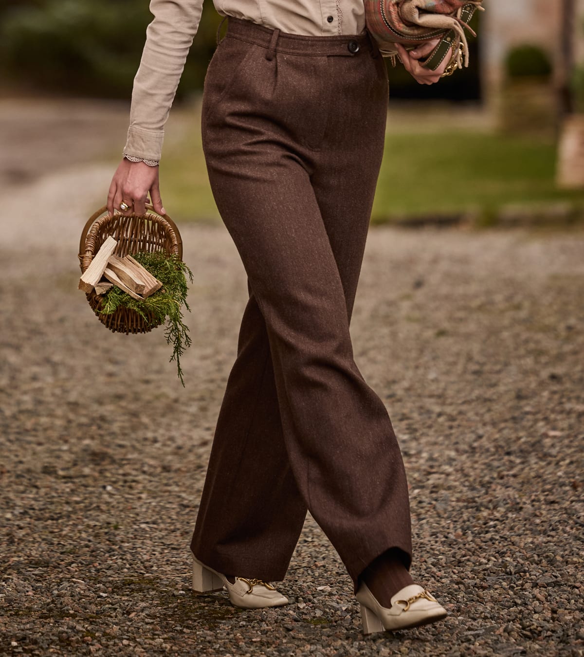 Person wearing brown wide-leg trousers, a cream shirt and loafers, holding a woven basket outdoors