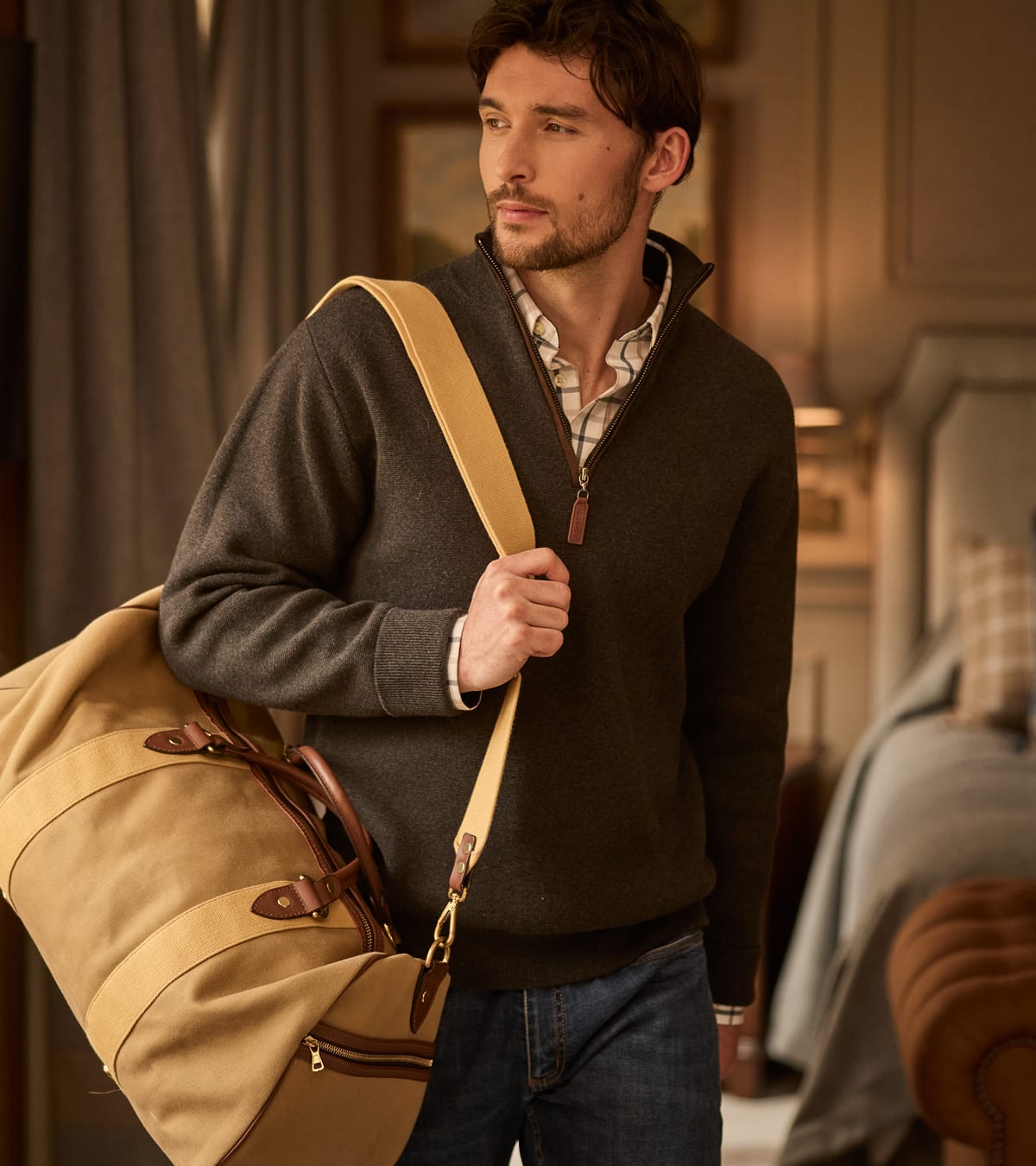 Man wearing a brown quarter-zip sweater and jeans, carrying a tan canvas duffel bag with leather handles.