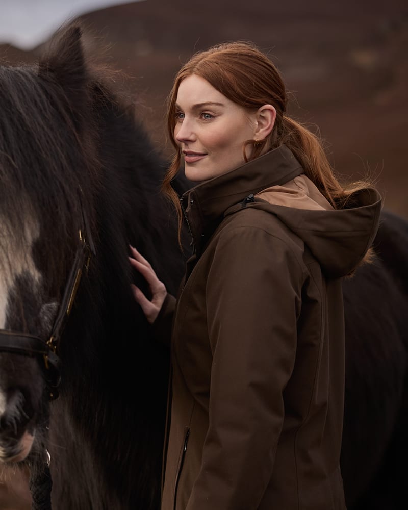 Red-haired woman wearing a brown hooded jacket standing beside a horse outdoors.