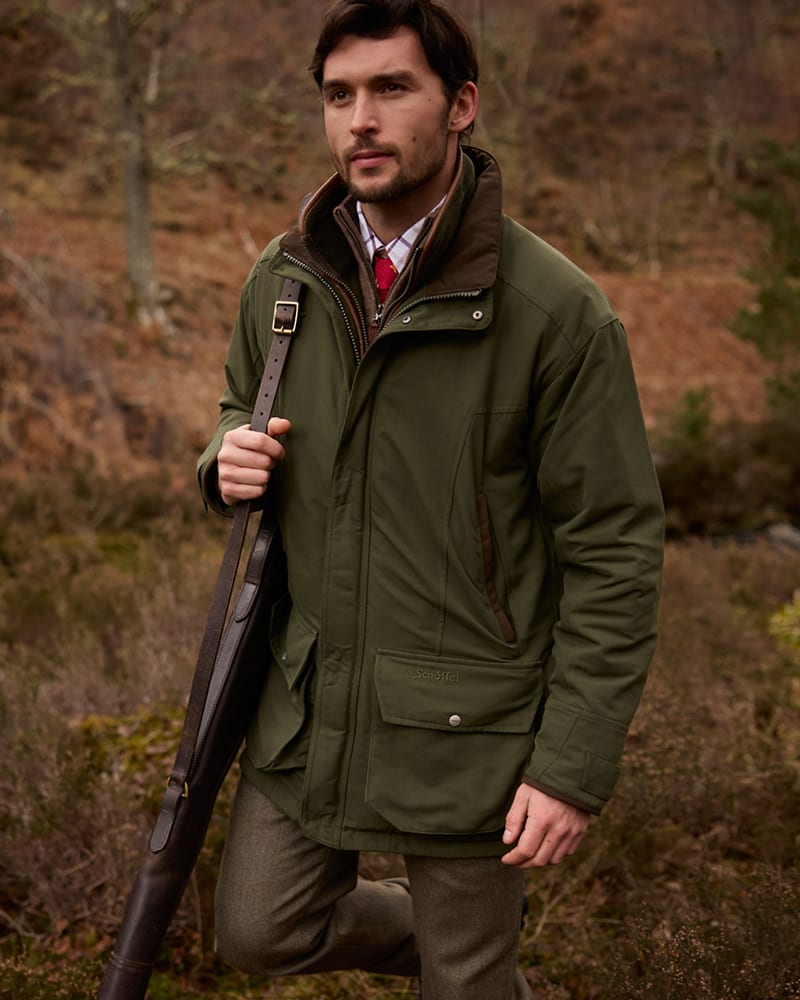 Man wearing an olive green hunting jacket with a brown collar, holding a shotgun in woodland.