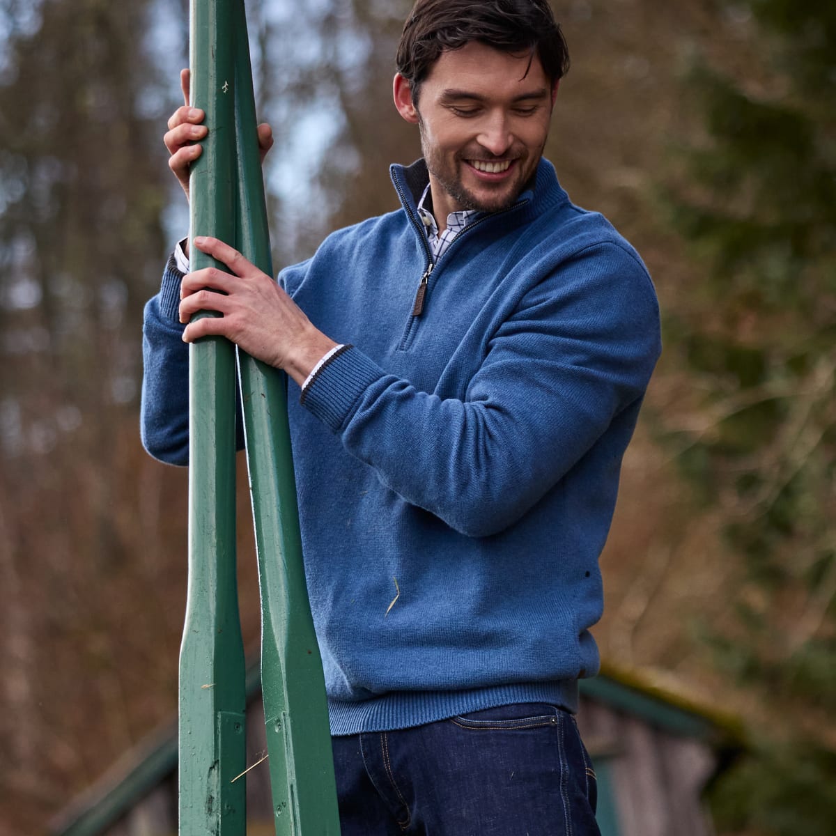 Man wearing a blue quarter-zip knit sweater and dark jeans outdoors among trees.