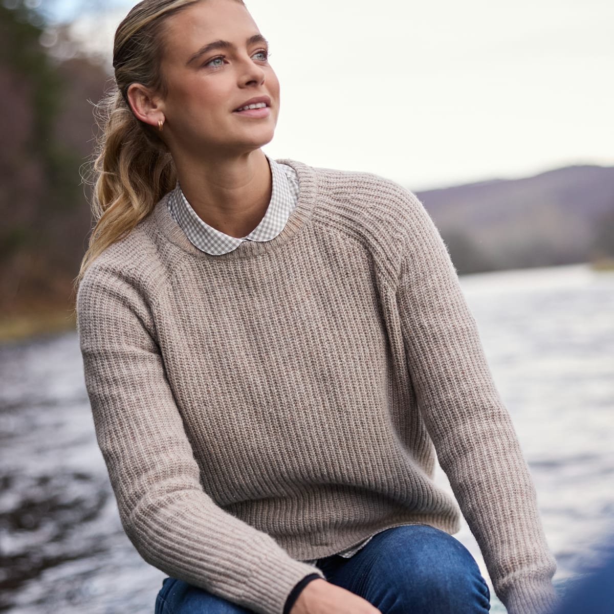 Woman wearing a beige ribbed knit sweater over a checked shirt beside a lake.