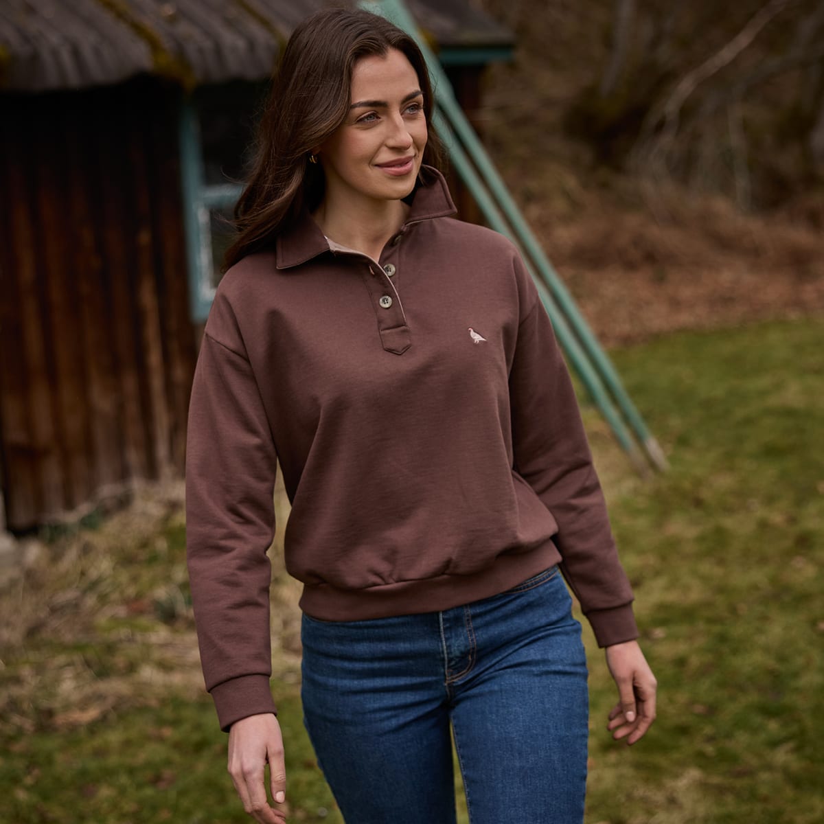 Woman wearing a brown long-sleeve polo top and blue jeans in a grassy field.