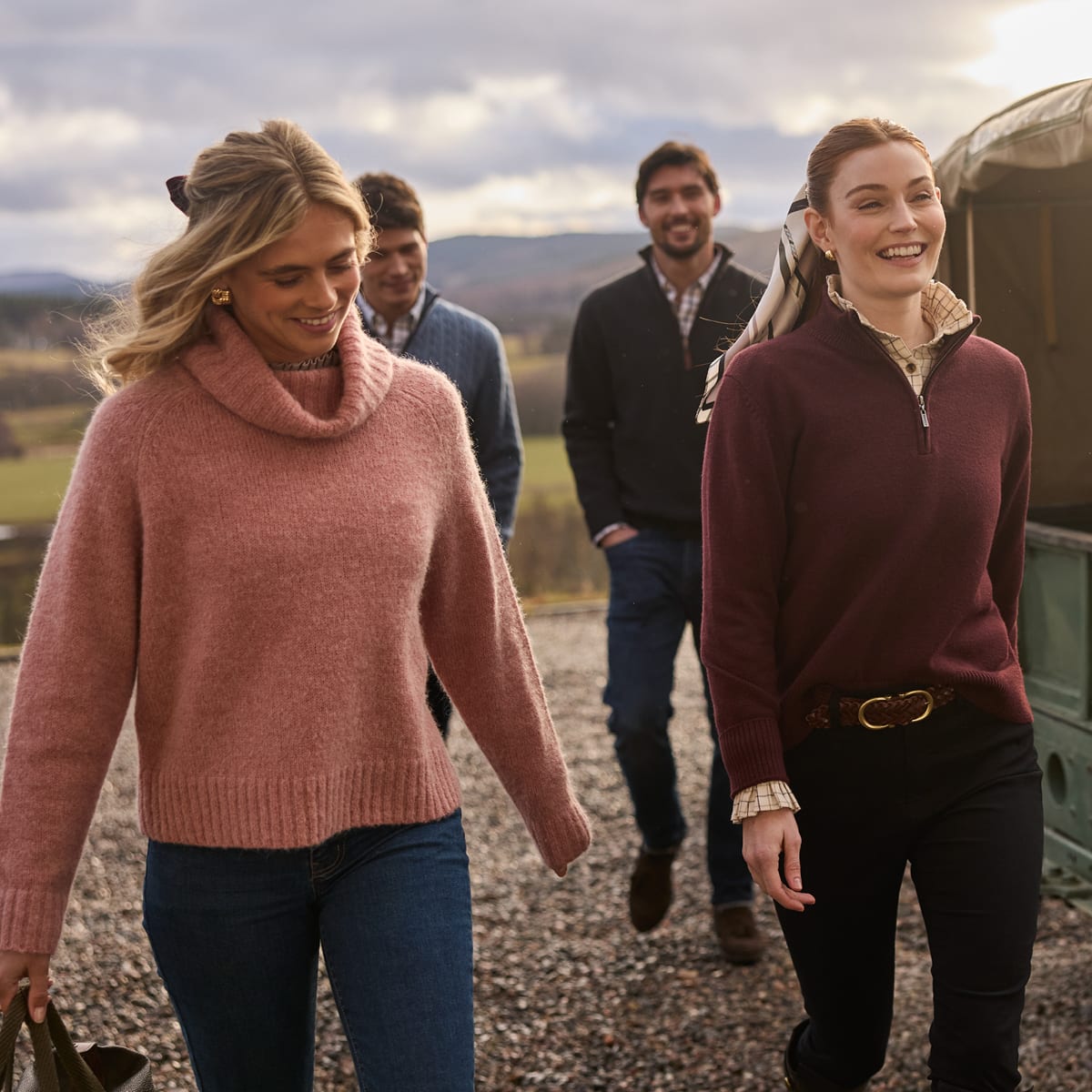 Four people wearing knit and quarter-zip sweaters in pink, burgundy, blue and navy, walking near a vintage green vehicle.