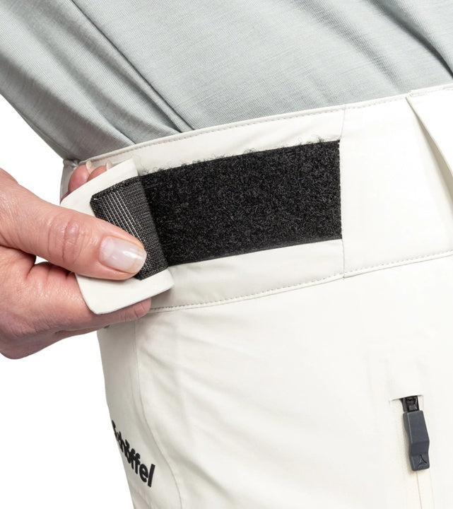 Close-up of a person adjusting a black strap on white ski trousers with a visible Schöffel logo.