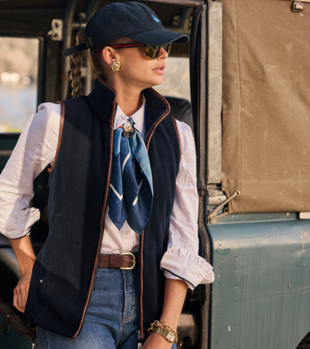 Person wearing a navy fleece gilet layered over a shirt and scarf, standing beside a vehicle outdoors.
