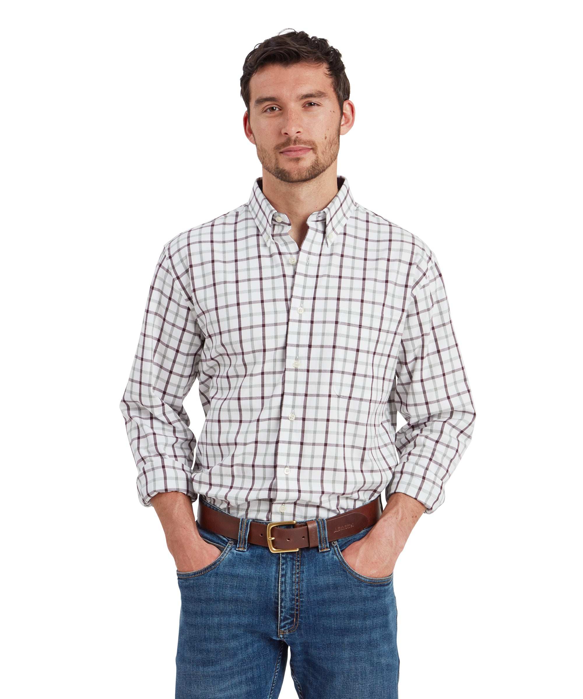 A man is wearing a Schöffel Brancaster Classic Shirt for Men in Red with a white base and a pattern of maroon and gray checks. The shirt is tucked into a pair of medium-wash jeans, secured with a brown leather belt. He is standing with his hands casually placed in his pockets, looking directly at the camera with a neutral expression. The shirt has a structured collar, and the sleeves are rolled up to the forearms.