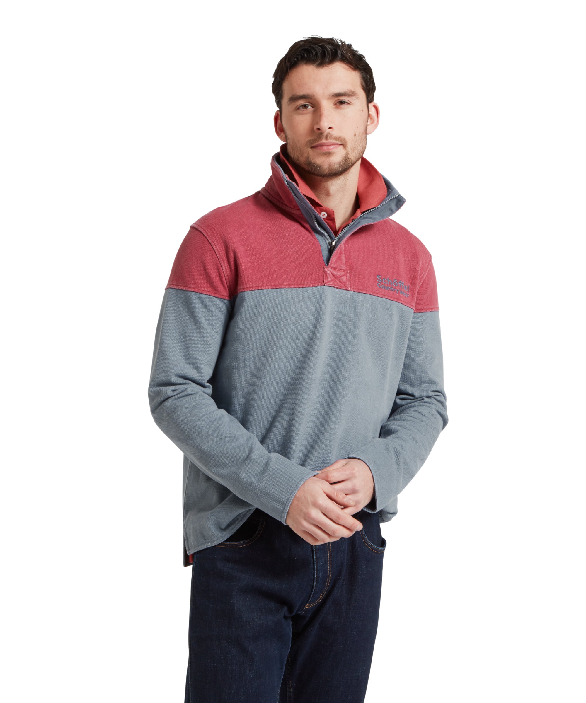 Half height image of a male model wearing a blue Schöffel sweatshirt with red shoulder and chest detailing and blue 'Schöffel countrywear' logo on the chest. It is paired with a red polo and dark denim jeans. He is standing in a relaxed pose looking towards the camera.