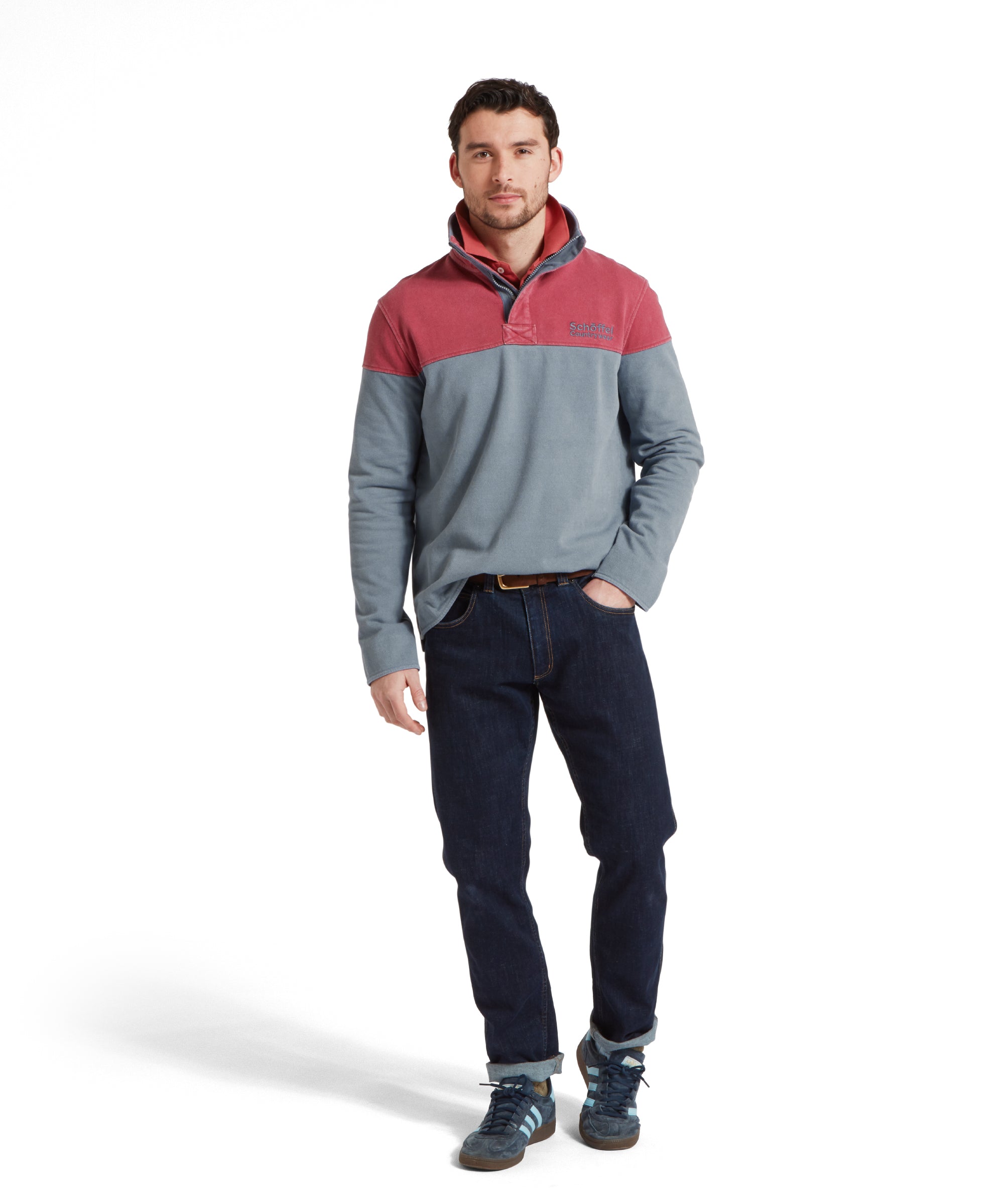 Full height image of a male model wearing a blue Schöffel sweatshirt with red shoulder and chest detailing and blue 'Schöffel countrywear' logo on the chest. It is paired with a red polo, navy trainers and dark denim jeans. He is standing in a relaxed pose looking towards the camera with one hand in his pocket.