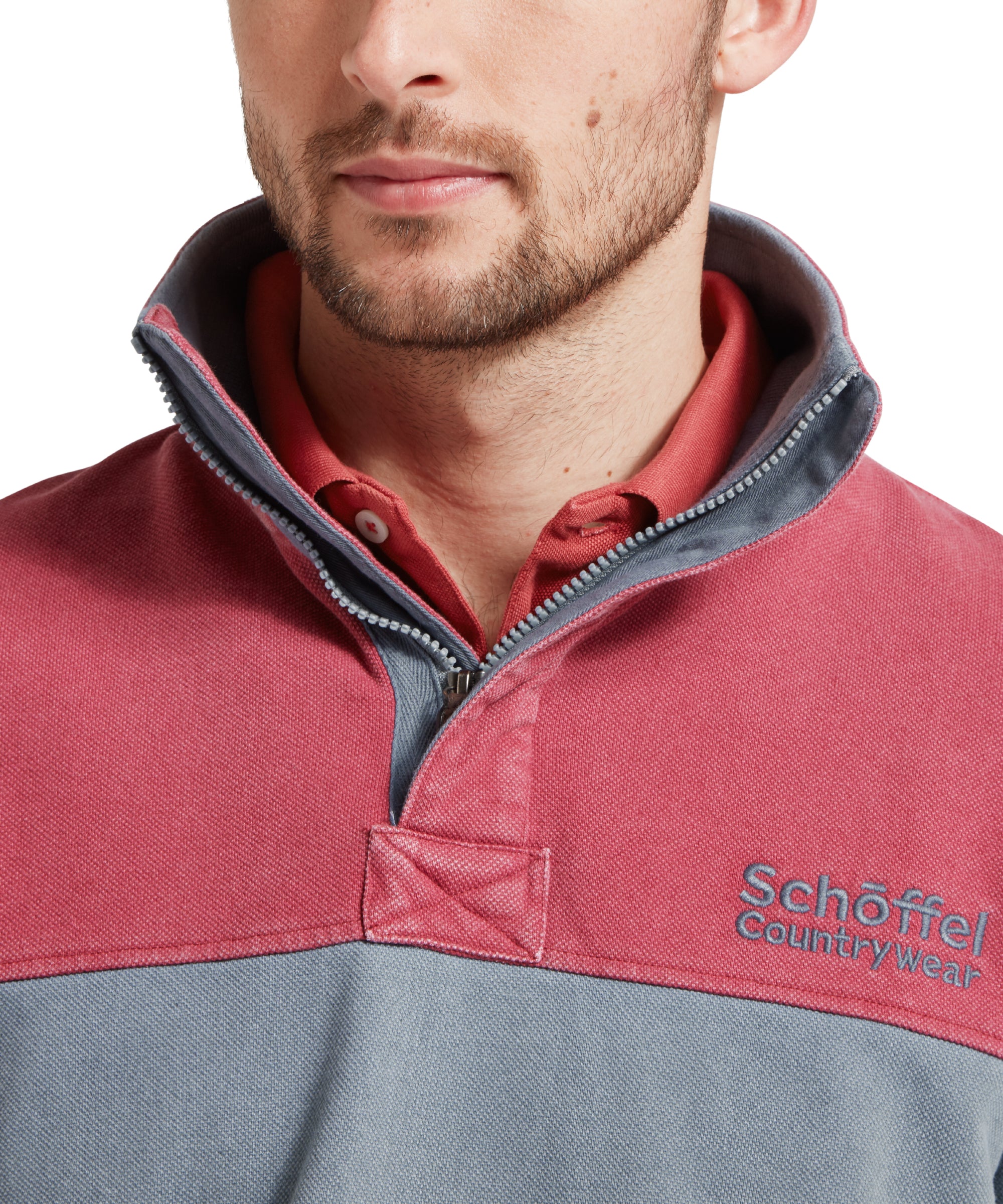 Close up image of a male model wearing a blue Schöffel sweatshirt with red shoulder and chest detailing and blue 'Schöffel countrywear' logo on the chest. It is paired with a red polo. Image highlights the quarter zip and collar detailing.