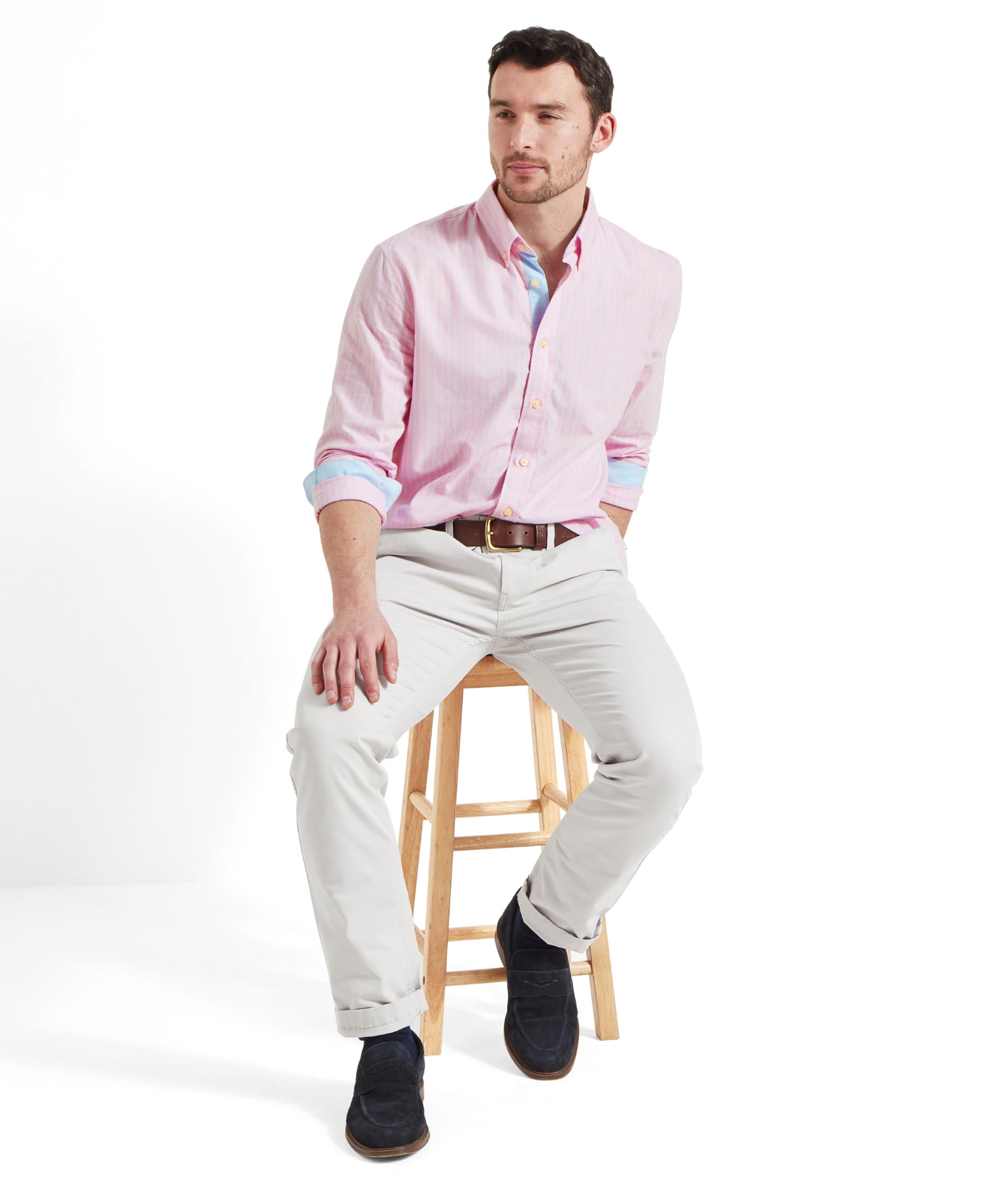 A man wears the Schöffel Holt Soft Oxford Tailored Shirt for Men in Pink Stripe whilst he perches on a wooden stool. He places his left hand on his knee and is glancing to the left. The shirt is worn with light beige trousers, tan leather belt and blue suede loafers.