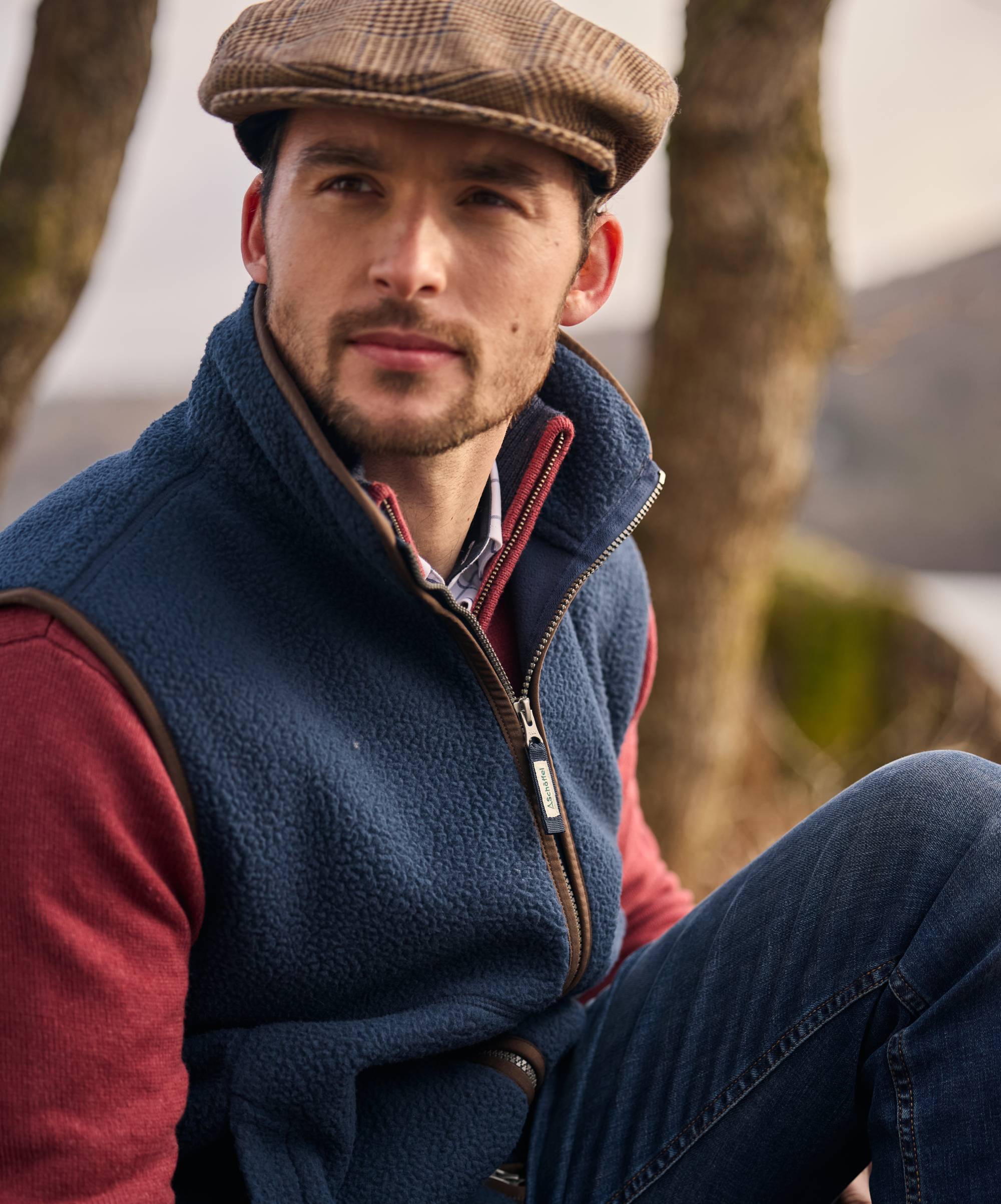 Man wearing a blue gilet over a red knitted jumper with a tweed cap, sitting outdoors.