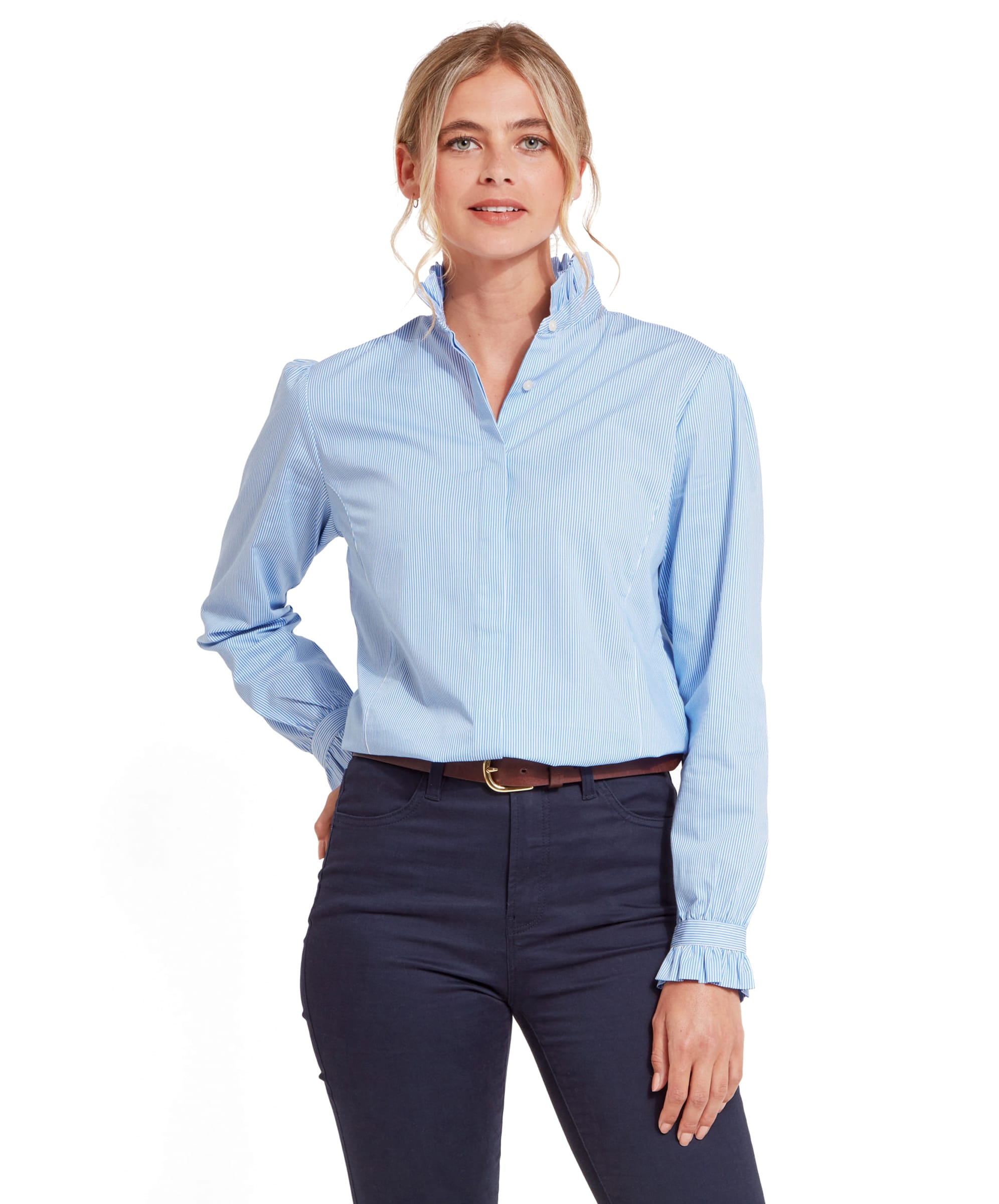 A woman wearing a Schöffel Fakenham Shirt for Womens in Blue with a ruffled collar and cuffs. She is standing with one hand on her hip, facing forward, and smiling. The shirt is paired with navy blue pants and a brown belt.
