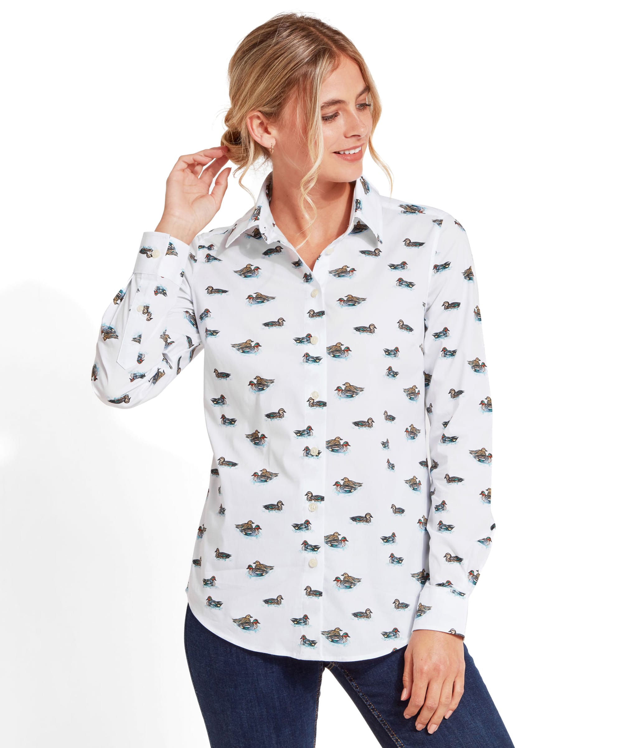 A woman wearing a Schöffel Norfolk Shirt for Womens in Blue with a print of small ducks. She has blonde hair tied back, smiling slightly, and is holding a strand of hair with one hand. The shirt is paired with dark jeans.