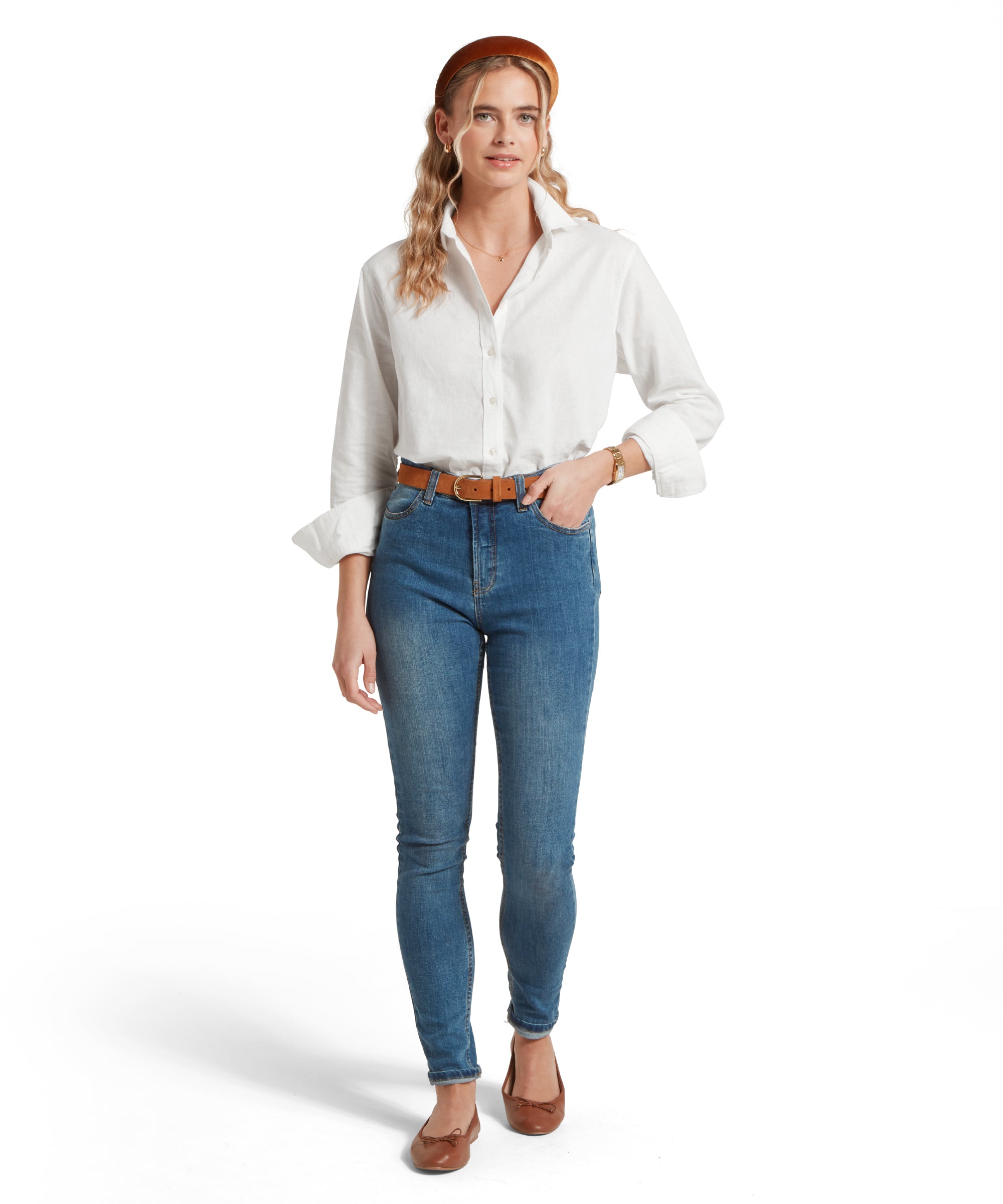 Full height image of a female model wearing a tucked in white linen Schöffel shirt, paired with mid denim jeans, tan pumps and tan leather belt. She is standing in a relaxed pose with one hand in her pocket.