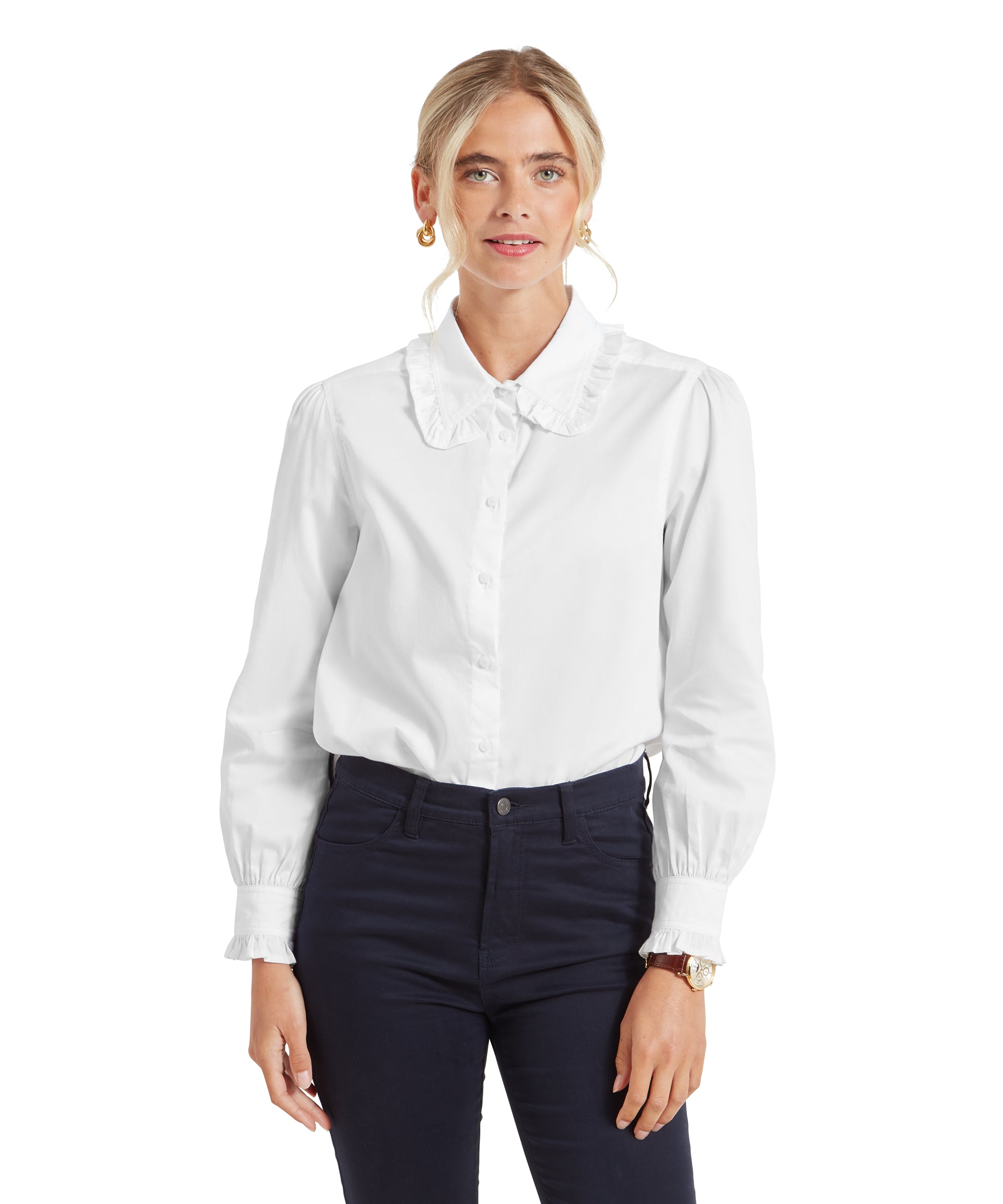 A woman is wearing a Schöffel Somerleyton Shirt for Women in White with a ruffled collar and long puff sleeves. She pairs it with dark navy skinny pants. Her blonde hair is styled back, and she is wearing gold hoop earrings. She stands facing forward with a slight smile.