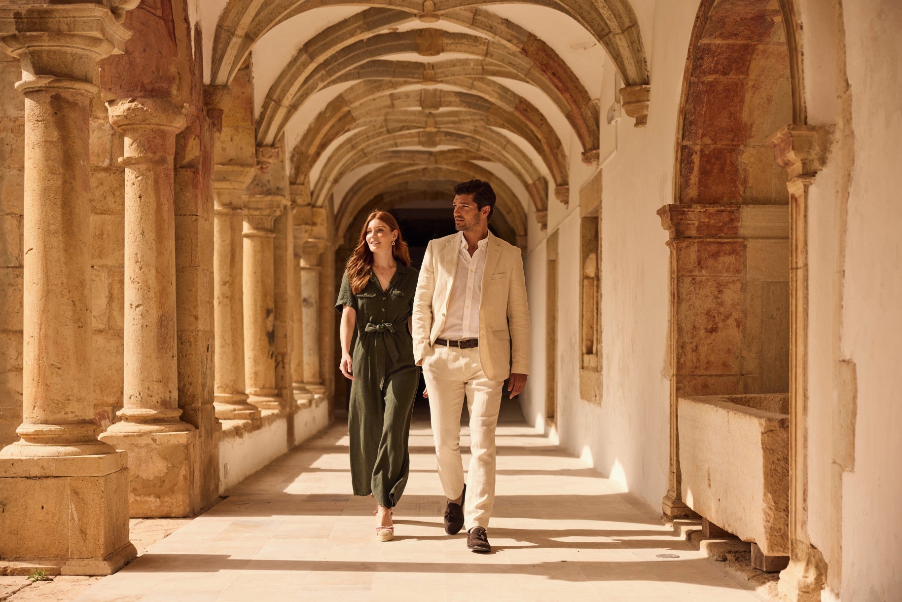 A couple walks side by side through a sunlit stone corridor with arches and columns, dressed in elegant, neutral-toned outfits, creating a warm and serene atmosphere.