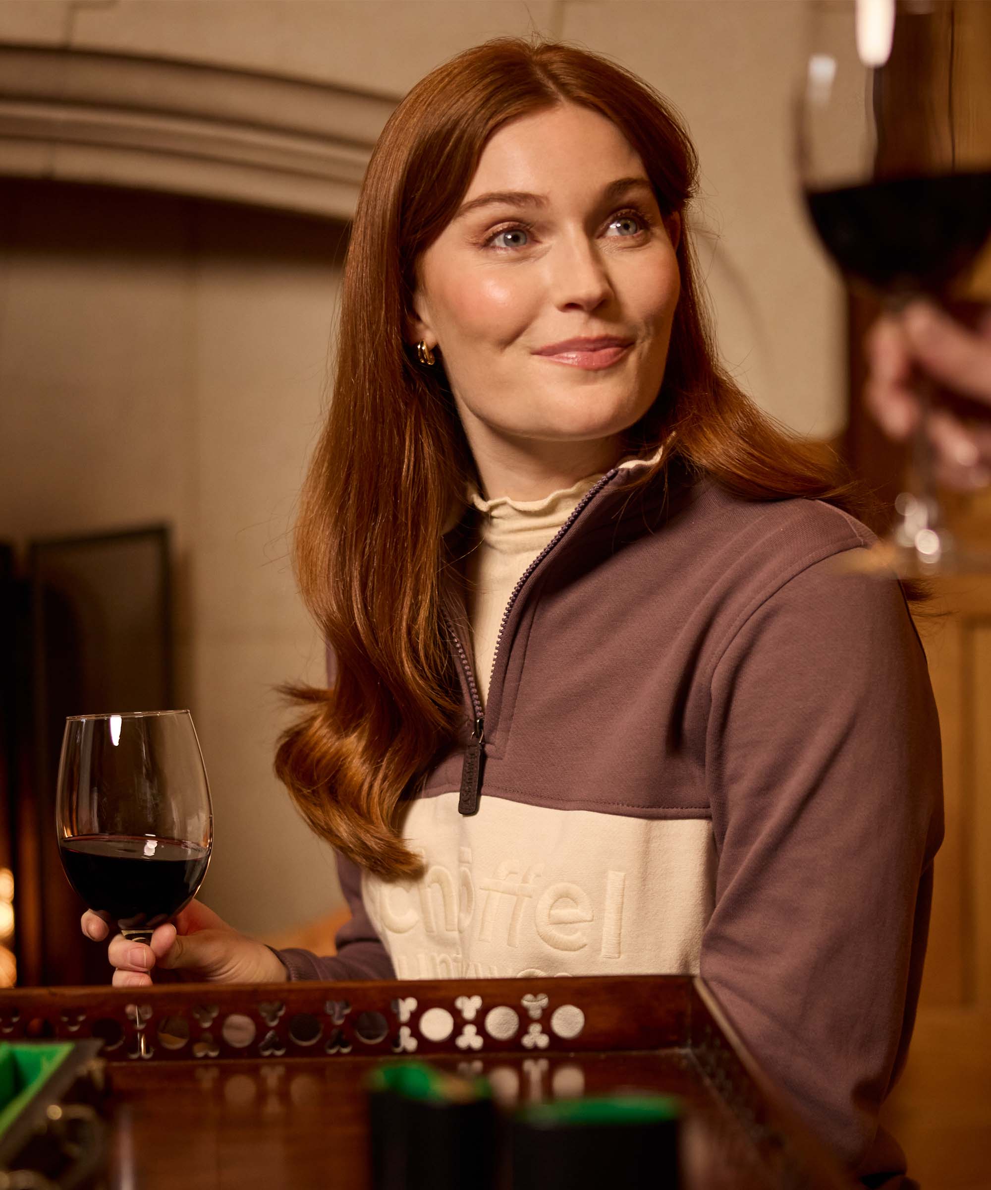 Woman enjoying a glass of red wine wearing a brown and oatmeal half-zip sweatshirt with embroidered Schöffel Countrywear branding, styled over a ruffle-neck top