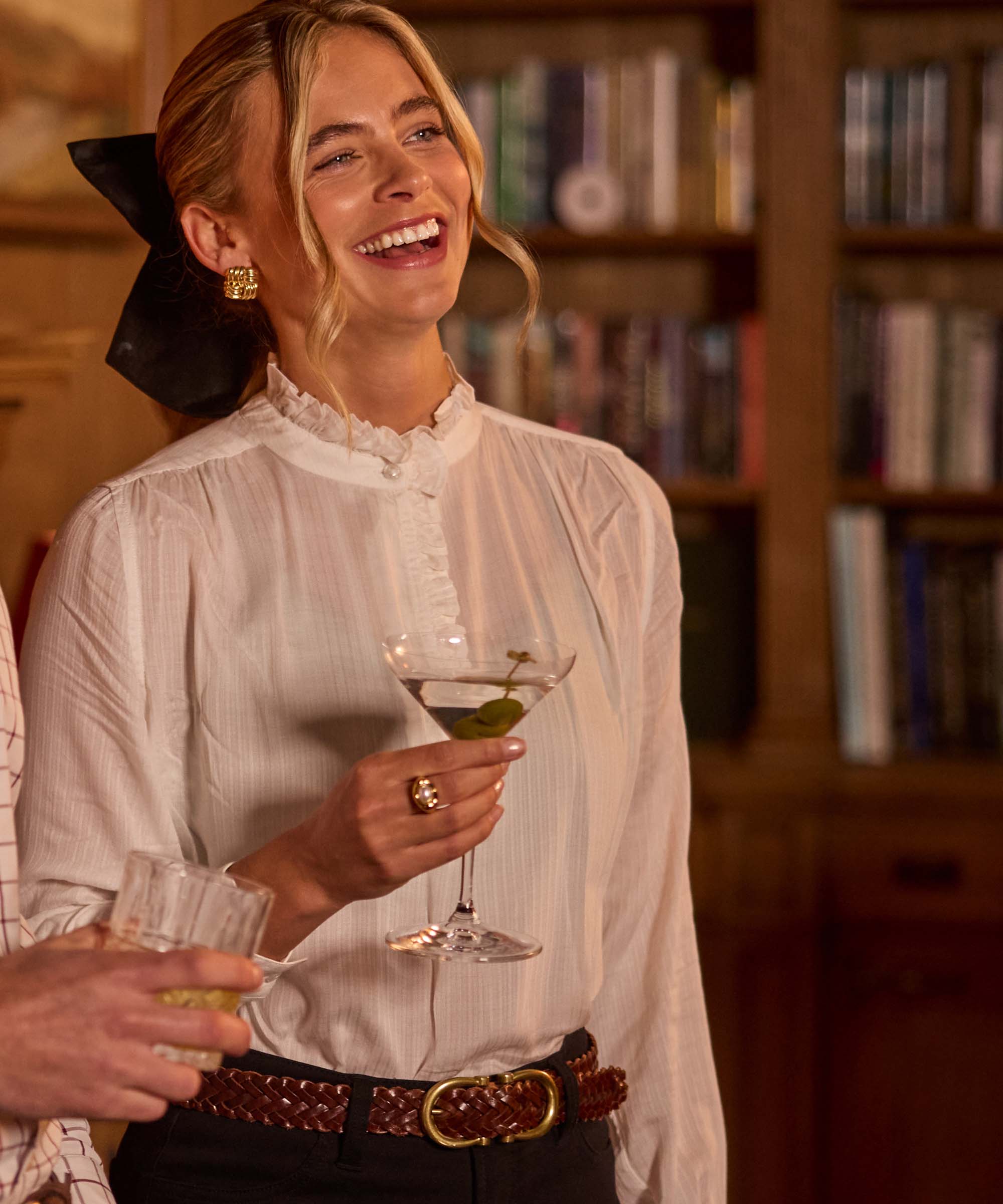 Woman laughing while holding a martini, wearing a soft white ruffle-neck blouse with a woven leather belt – relaxed elegance from Schöffel Country