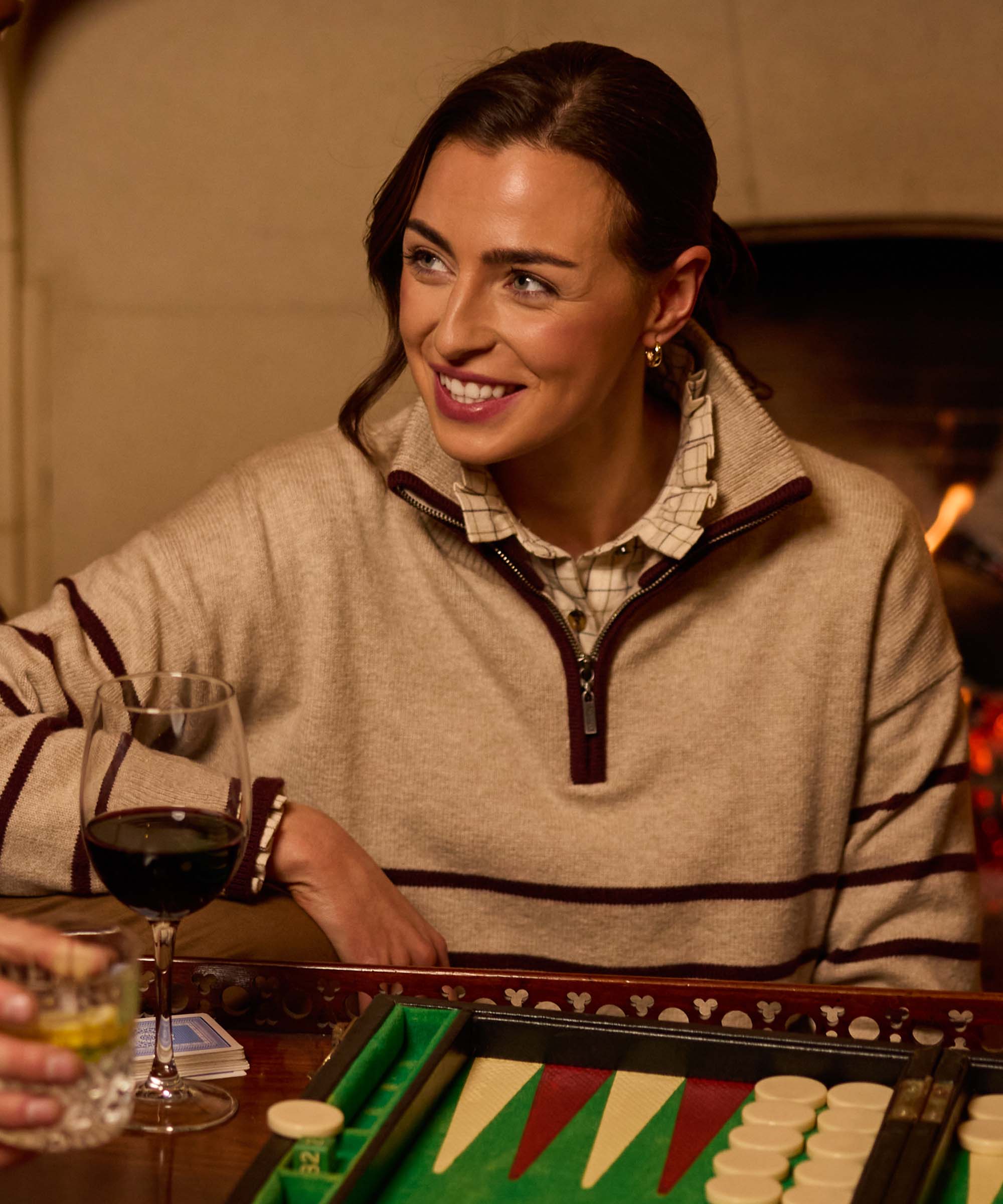 Woman smiling over a backgammon board holding a glass of red wine, wearing a beige and burgundy striped quarter-zip jumper over a check shirt