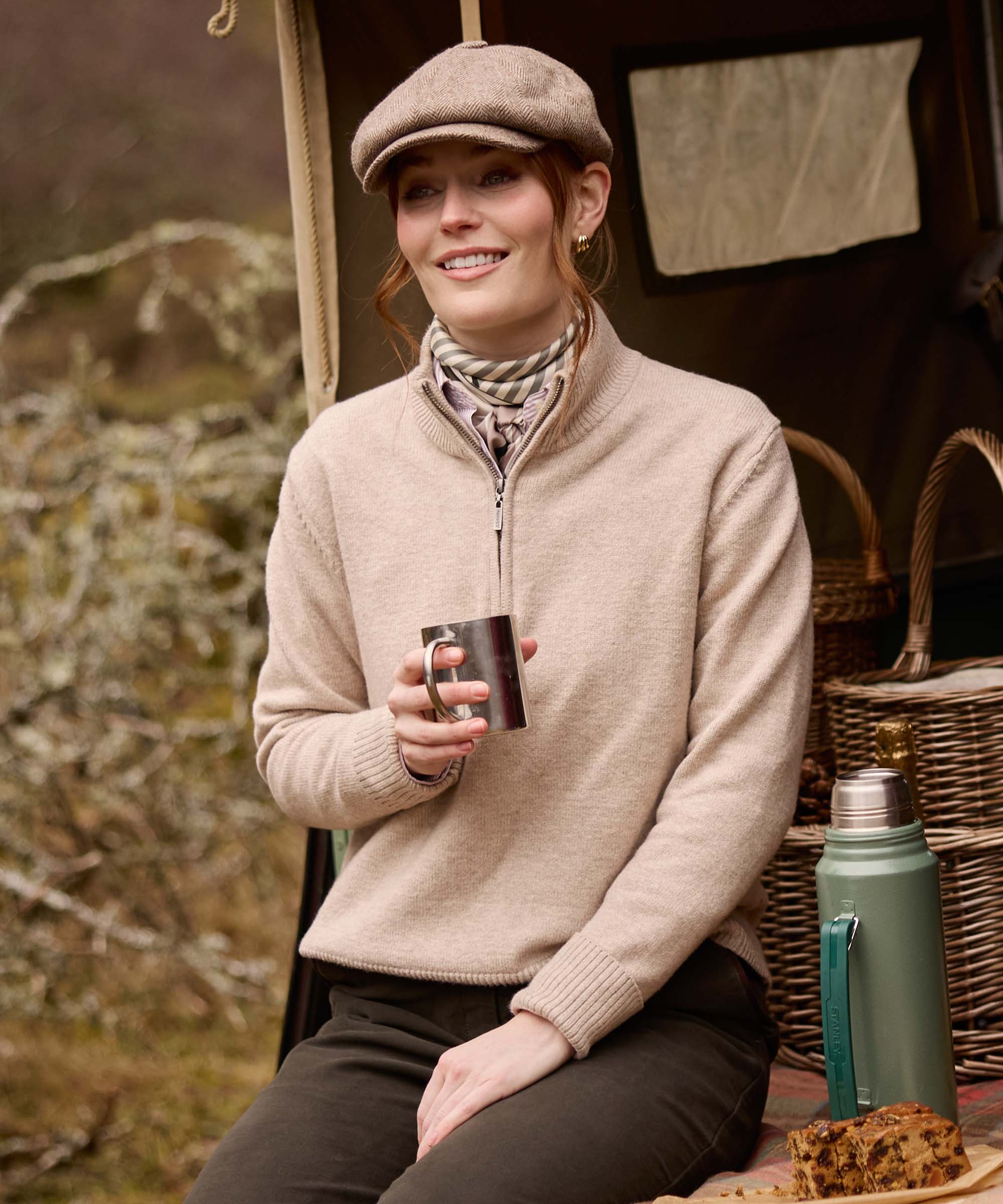 Woman in an oatmeal quarter-zip jumper and herringbone flat cap, holding a steel mug beside a wicker picnic hamper