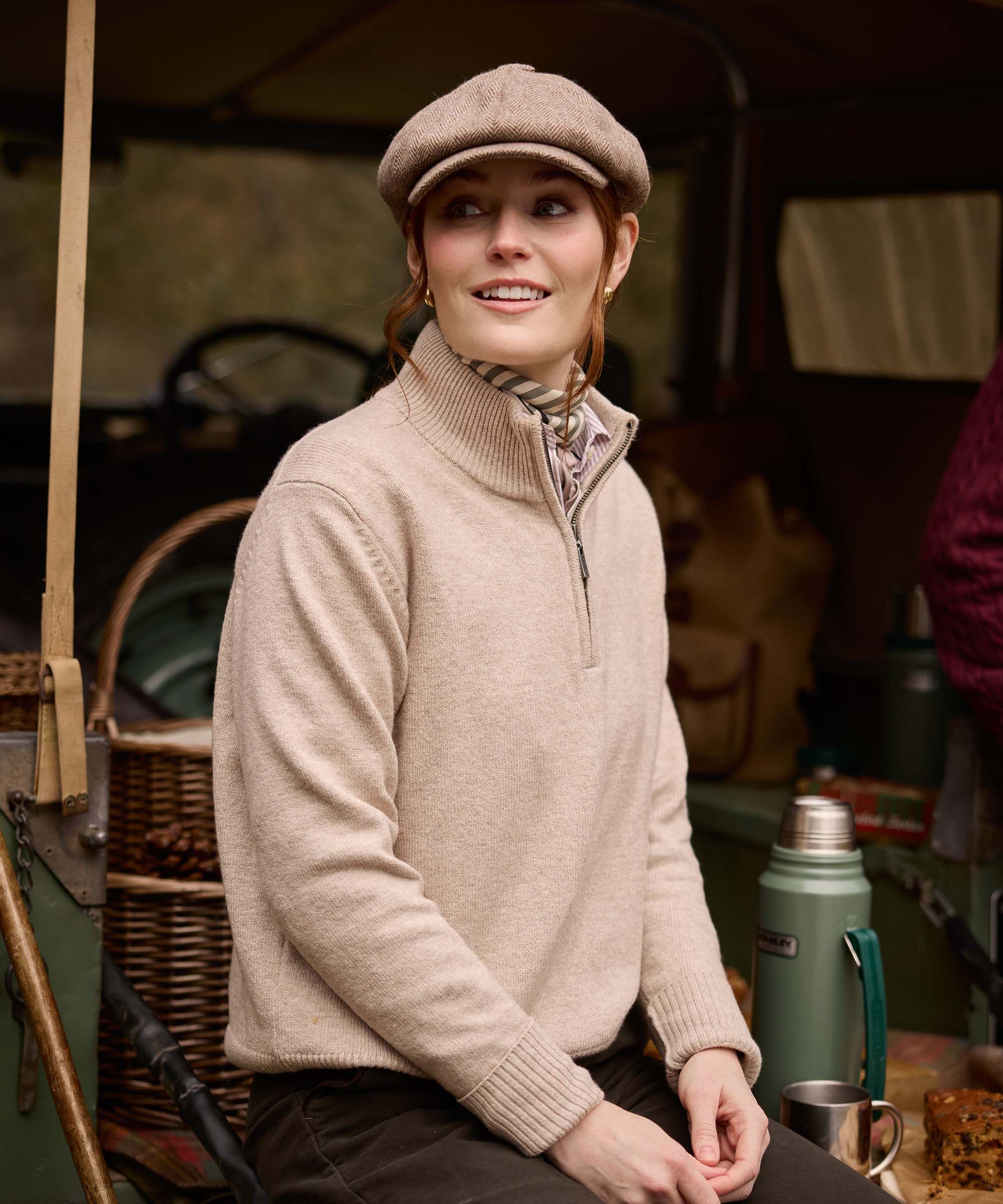 Woman wearing a herringbone flat cap and oatmeal quarter-zip jumper, smiling beside a vintage Land Rover shooting brake