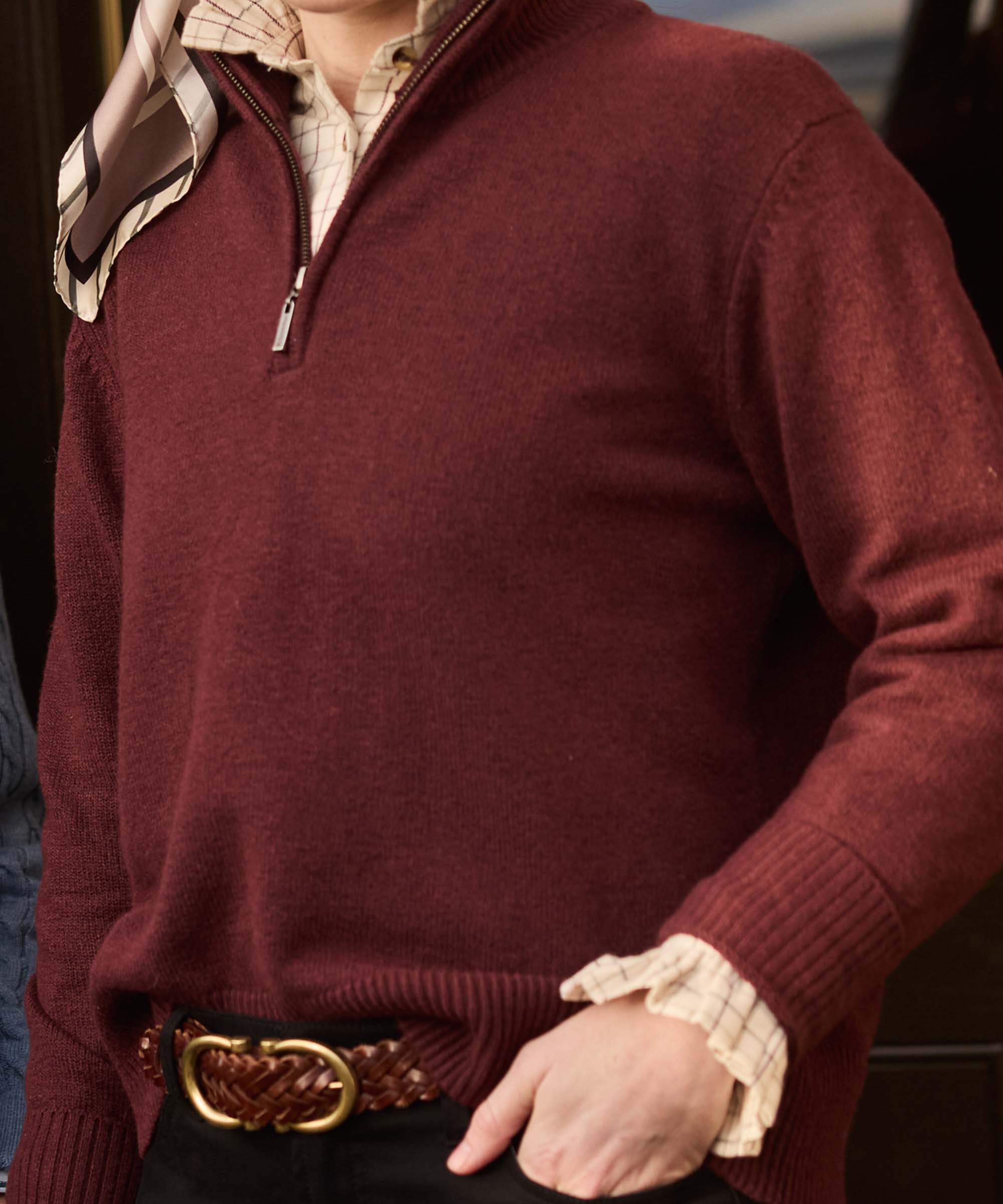Close-up of a burgundy quarter-zip jumper with ribbed cuffs, layered over a checked shirt and woven leather belt with brass buckle