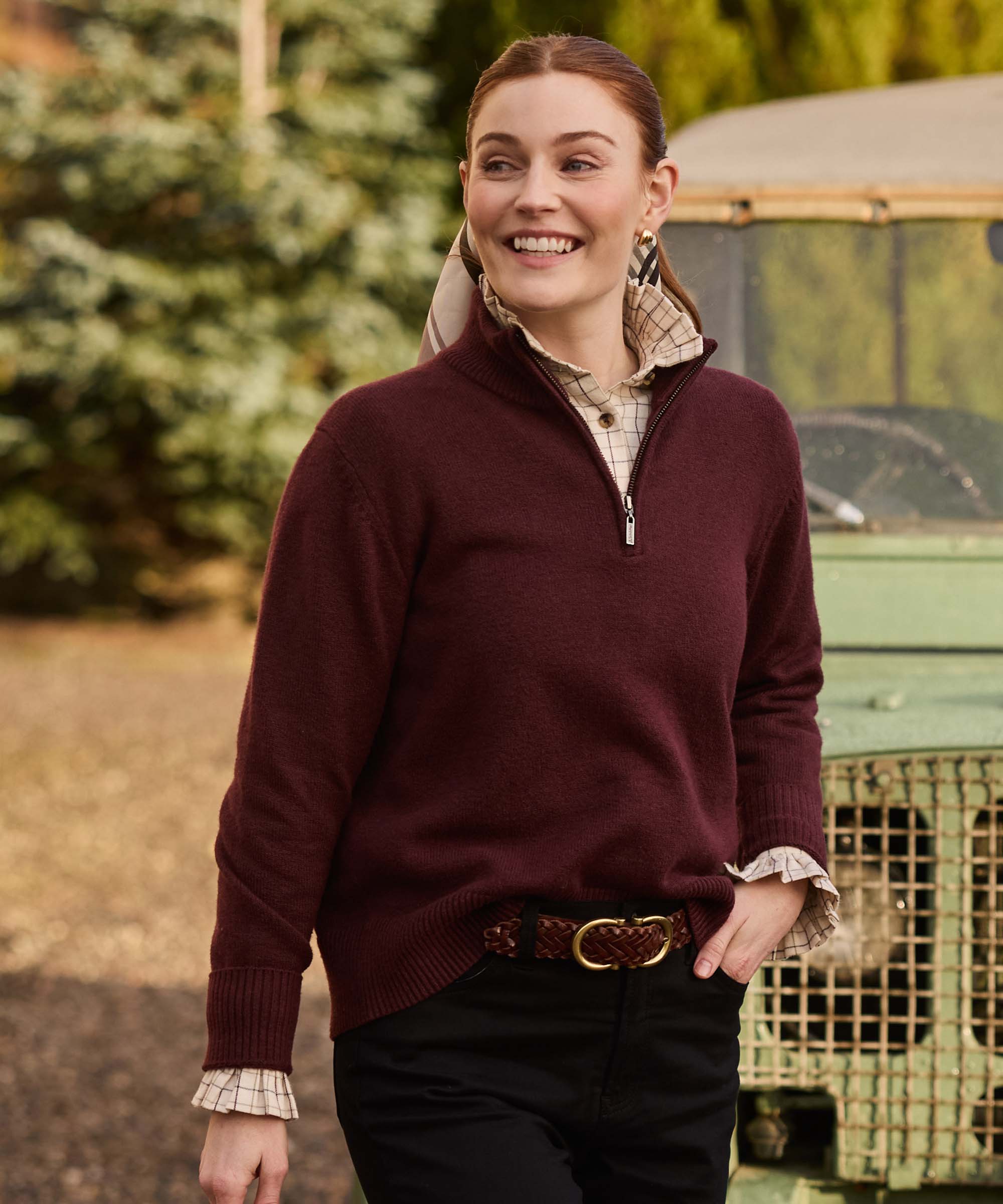 Woman walking outdoors in a burgundy quarter-zip jumper layered over a check shirt, finished with a woven leather belt