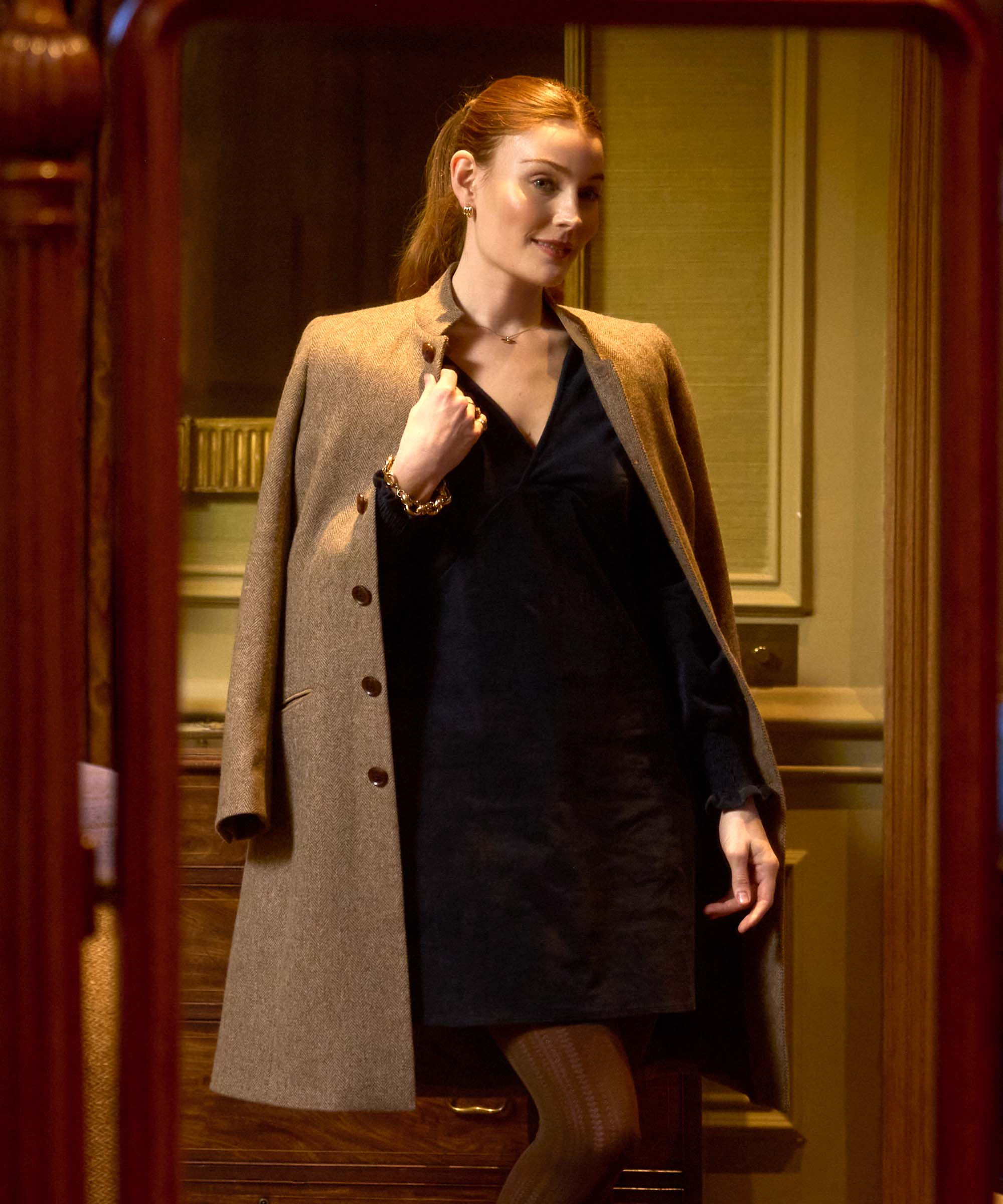 Woman in a tailored camel wool coat worn open over a navy velvet dress, styled with gold jewellery for elegant evening countryside style