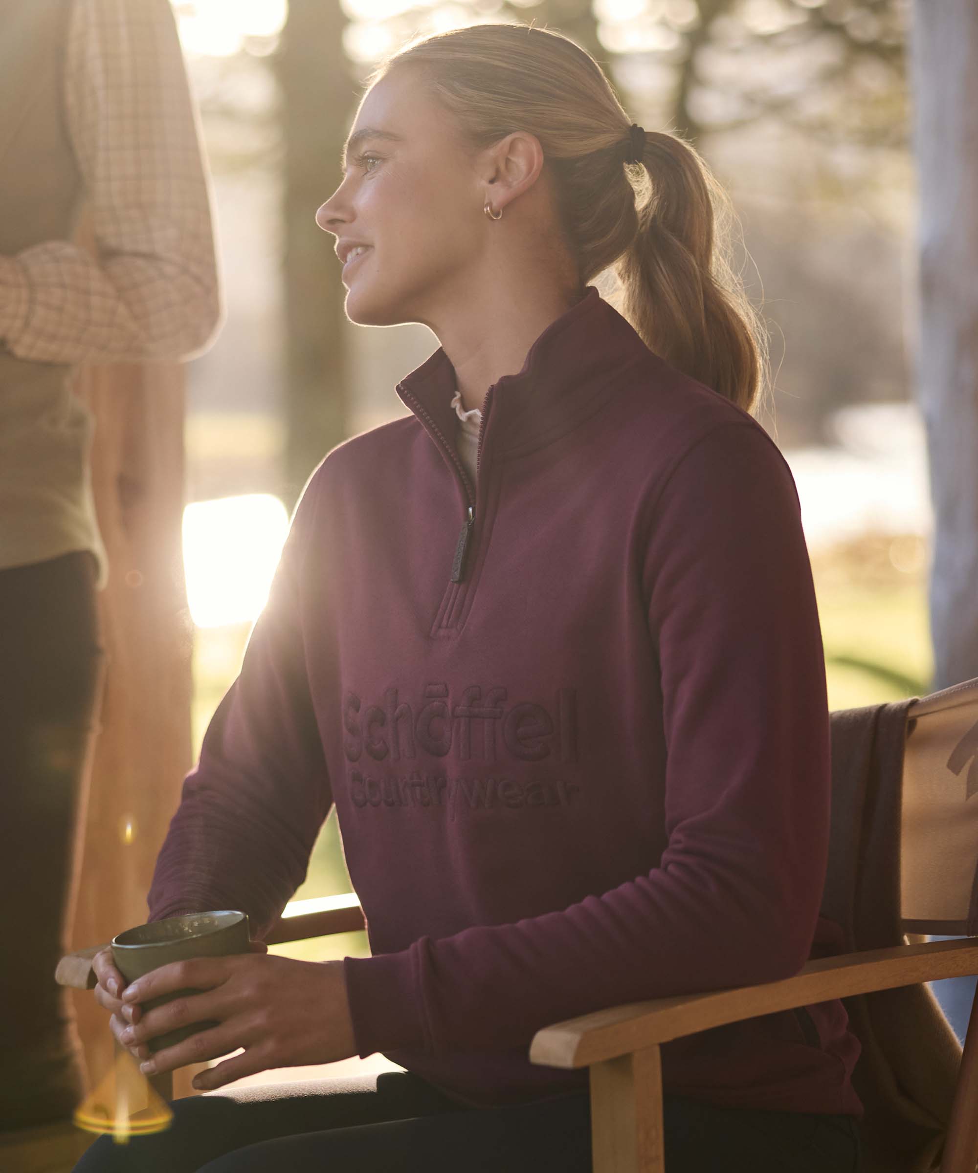 Woman in a burgundy quarter-zip jumper embroidered with the Schöffel Countrywear logo, relaxing outdoors in warm evening light