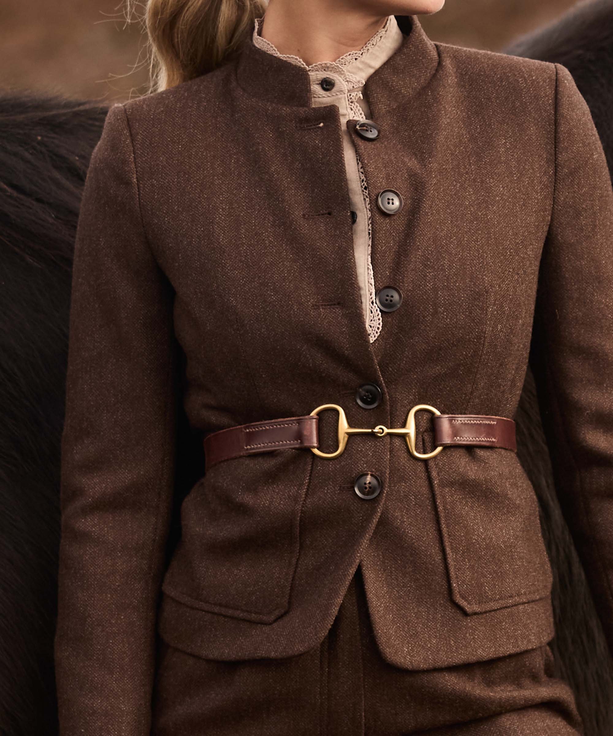 Close-up of a brown herringbone tweed jacket with stand collar and brass snaffle-bit belt detail, styled over a lace-trimmed blouse