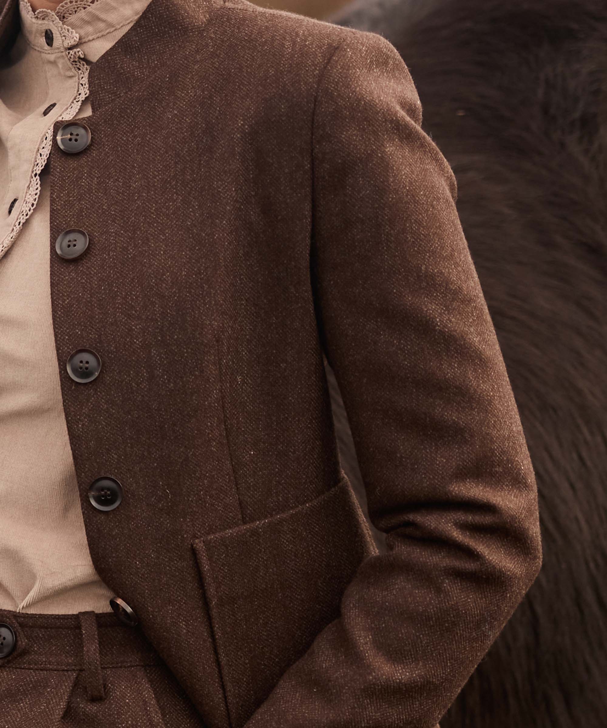 Close-up of a brown herringbone tweed jacket with stand collar and horn-effect buttons, layered over a taupe lace-edged blouse
