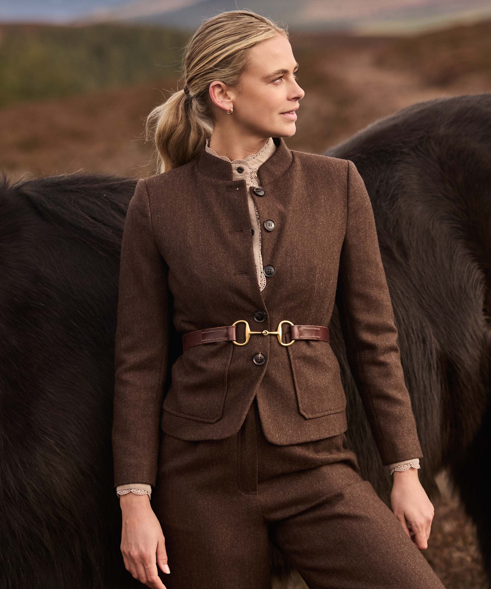 Woman standing beside a Highland pony wearing a brown herringbone tweed jacket with stand collar, cinched with a brown leather snaffle-bit belt, styled over a lace-trimmed blouse