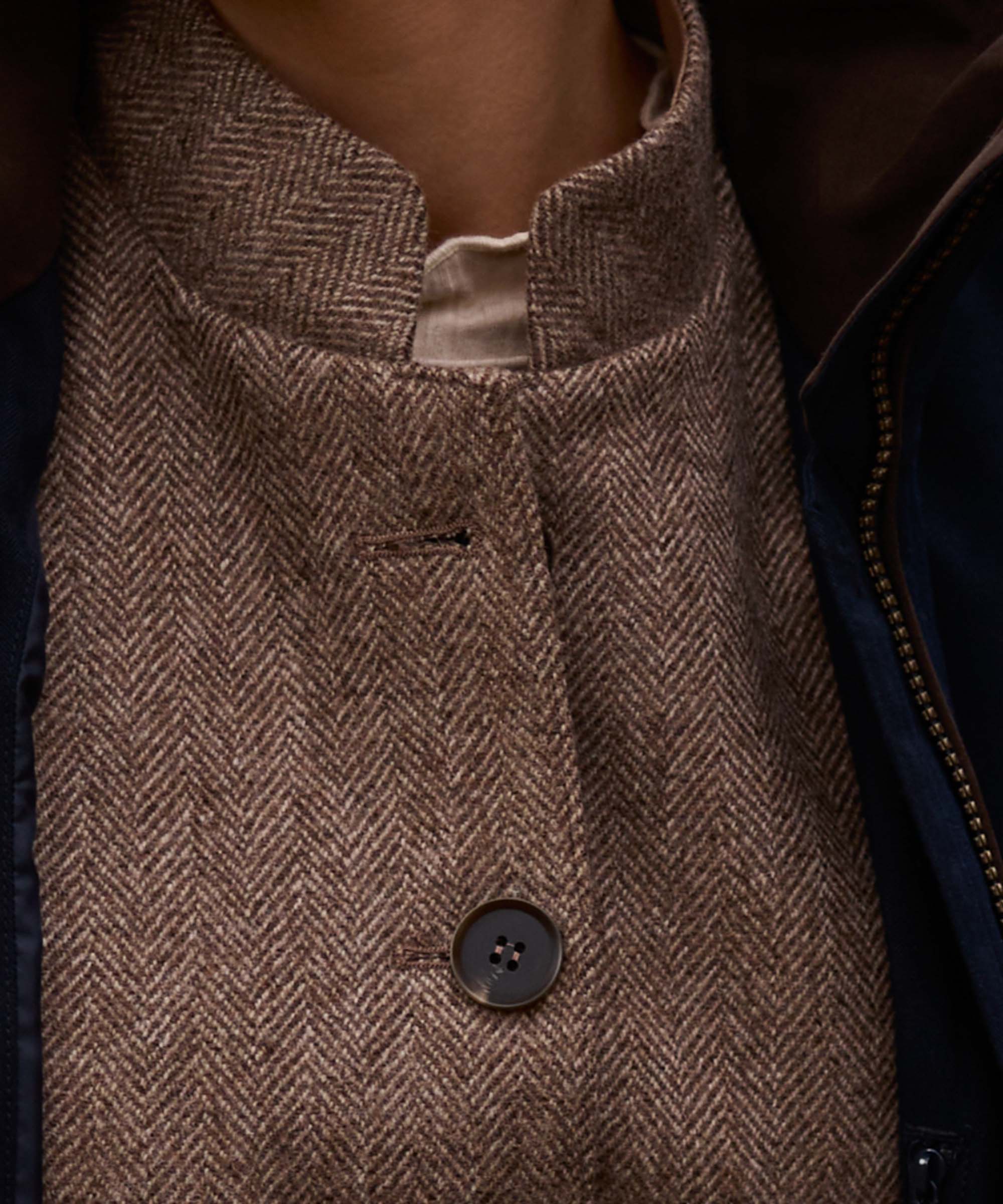 Close-up of a brown and beige herringbone patterned tweed coat with a high stand collar and a dark button, worn over a dark navy jacket.
