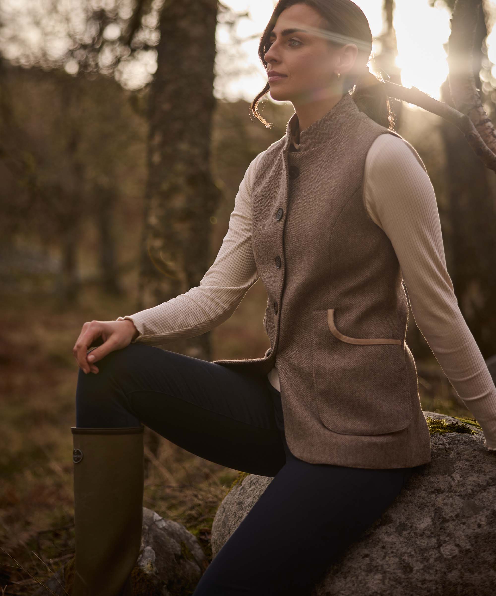 Woman seated outdoors in golden light wearing a taupe herringbone tweed gilet with suede-trimmed pockets, layered over a cream ribbed funnel-neck top