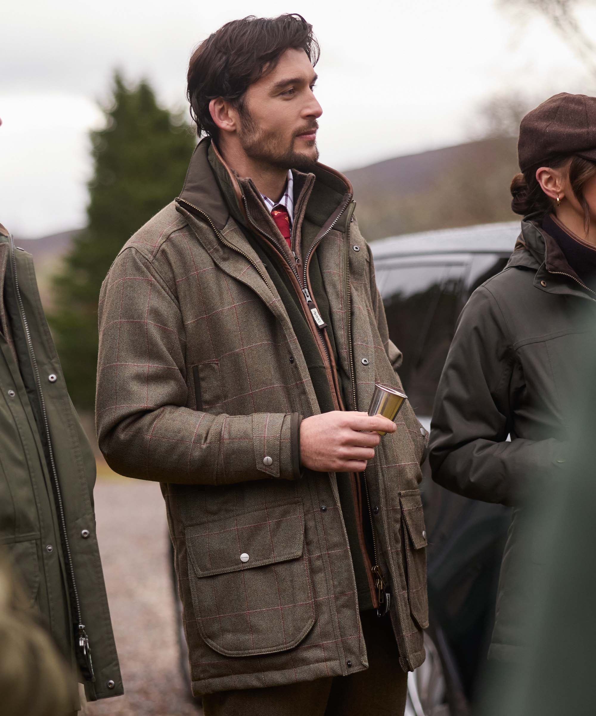 Man in an olive tweed shooting coat with leather-piped zip and multiple pockets, holding a hip flask cup while chatting outdoors